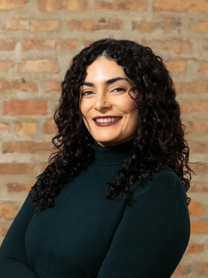 Woman wearing a brown turtleneck and necklace in front of a brick wall.