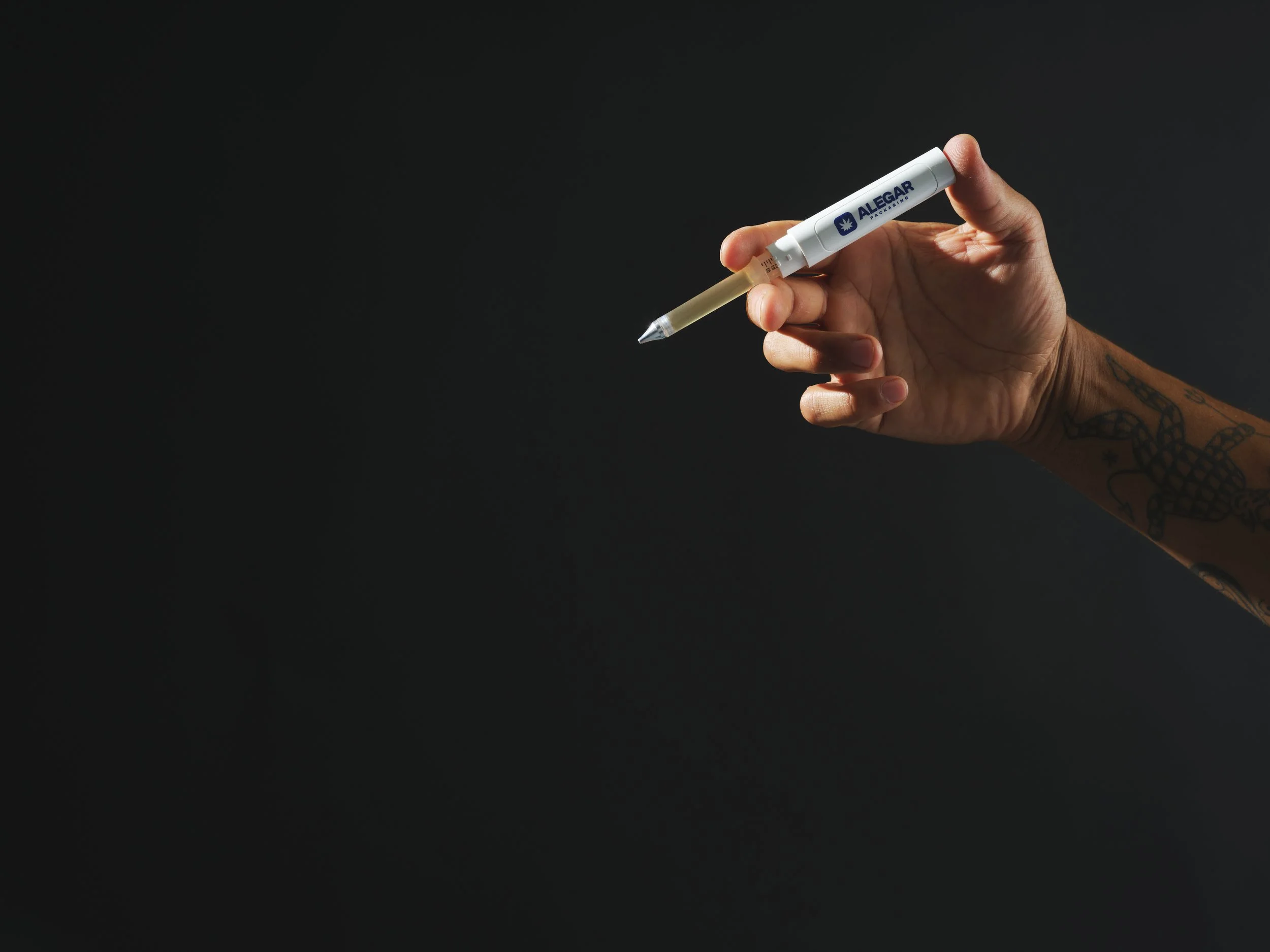 Close-up of a hand with tattoo holding a white marker with blue text reading 'ALEGAR PACKAGING' against a dark background.