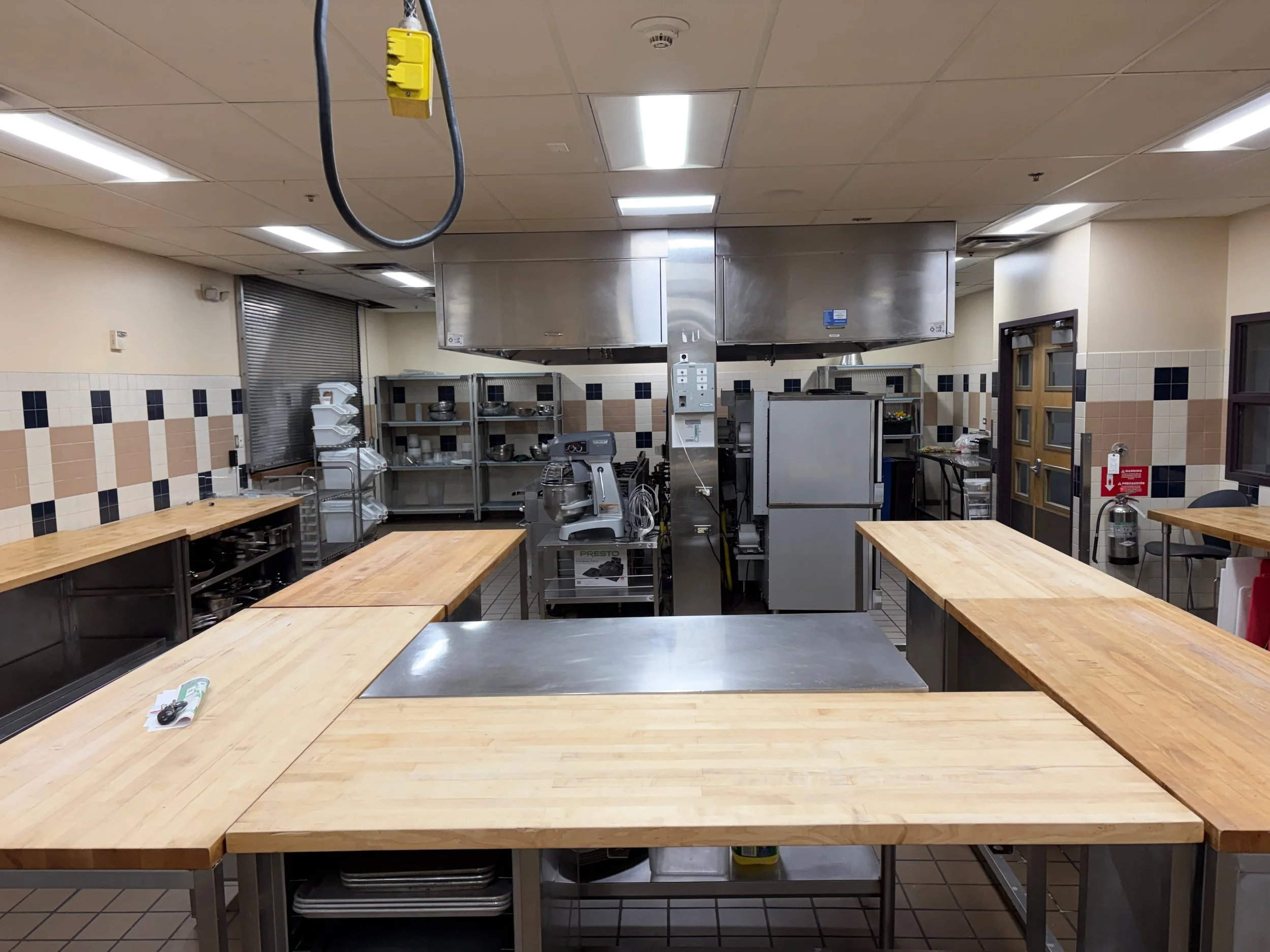 Empty commercial kitchen with wooden countertops, stainless steel appliances, and shelf with kitchenware, under fluorescent lighting.