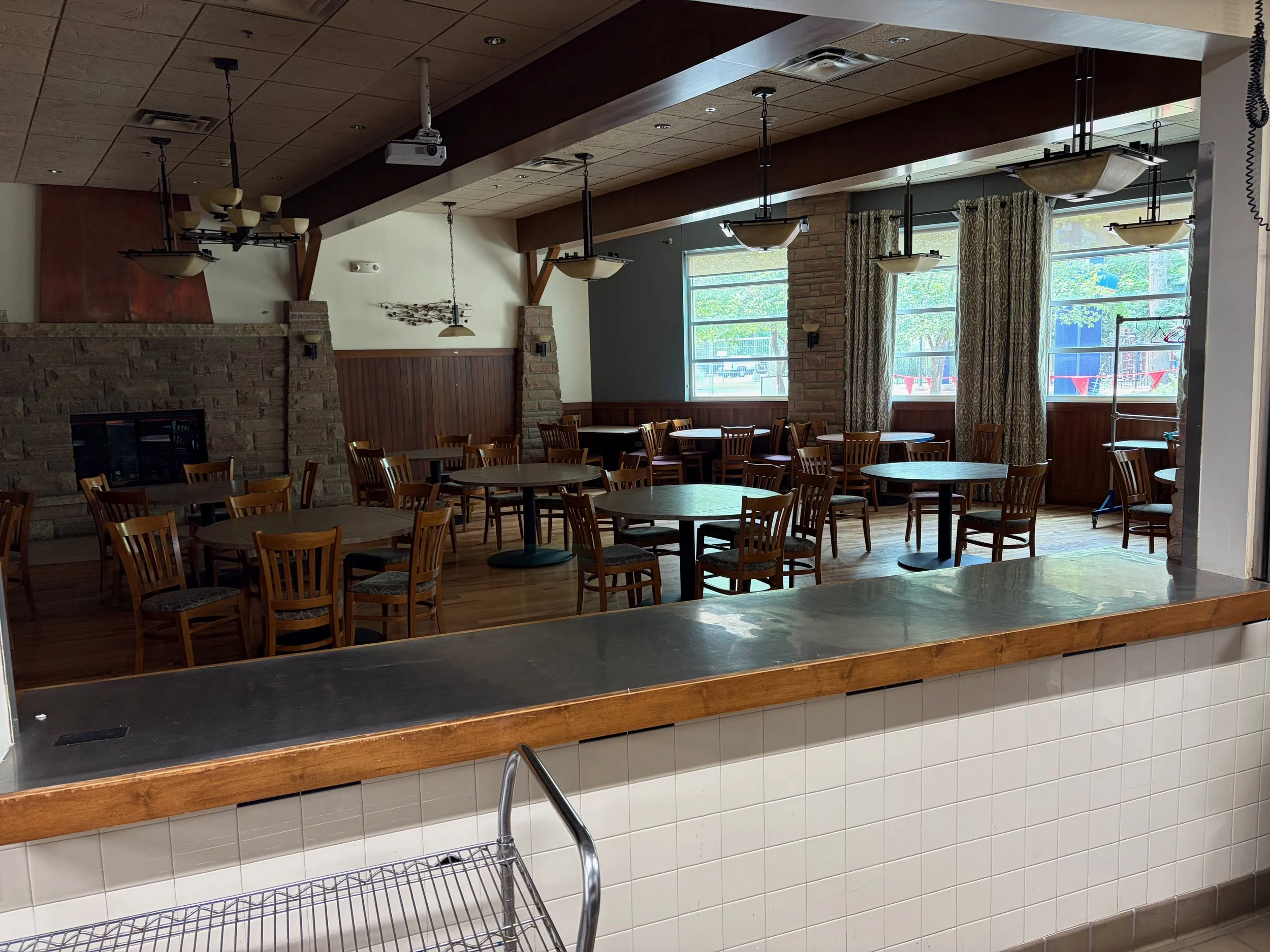 Interior of a restaurant or cafe with wooden chairs and tables, large windows with curtains, hanging light fixtures, and a brick and wood decor. A projection device is mounted on the ceiling.