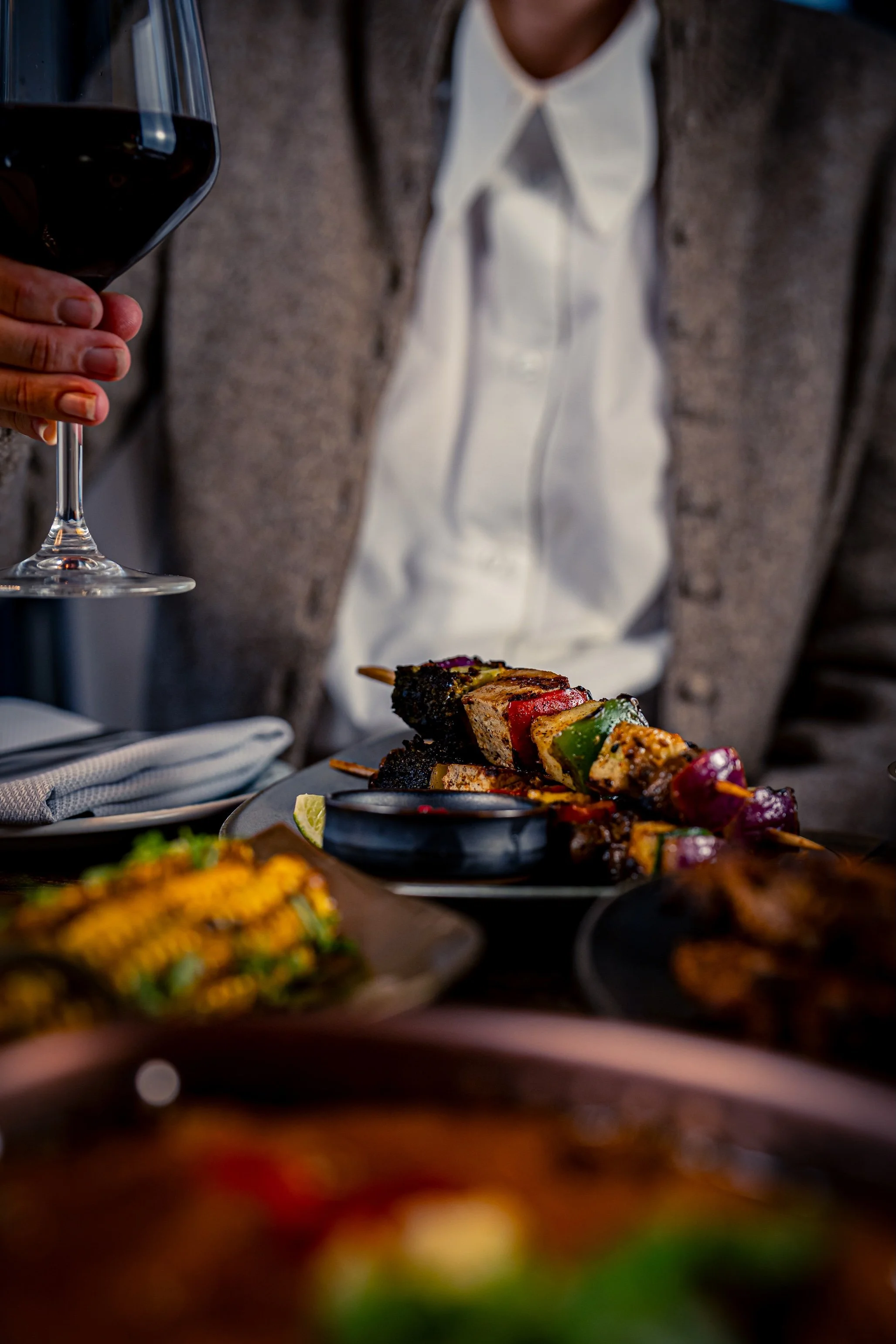 Person holding a glass of red wine with a plate of assorted grilled skewers and a bundle of grilled vegetables on a dining table.