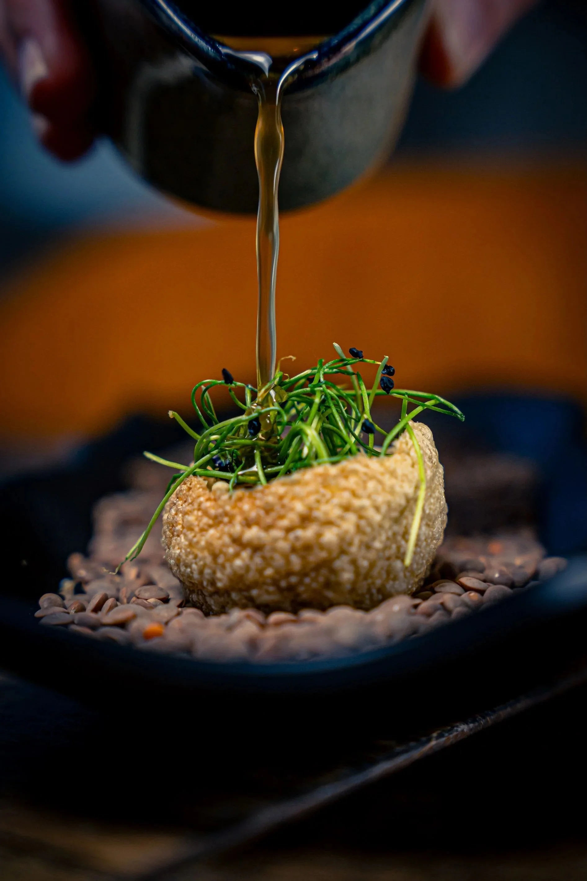 A breaded croquette topped with microgreens and black caviar, with a stream of sauce being poured over it, on a black tray.