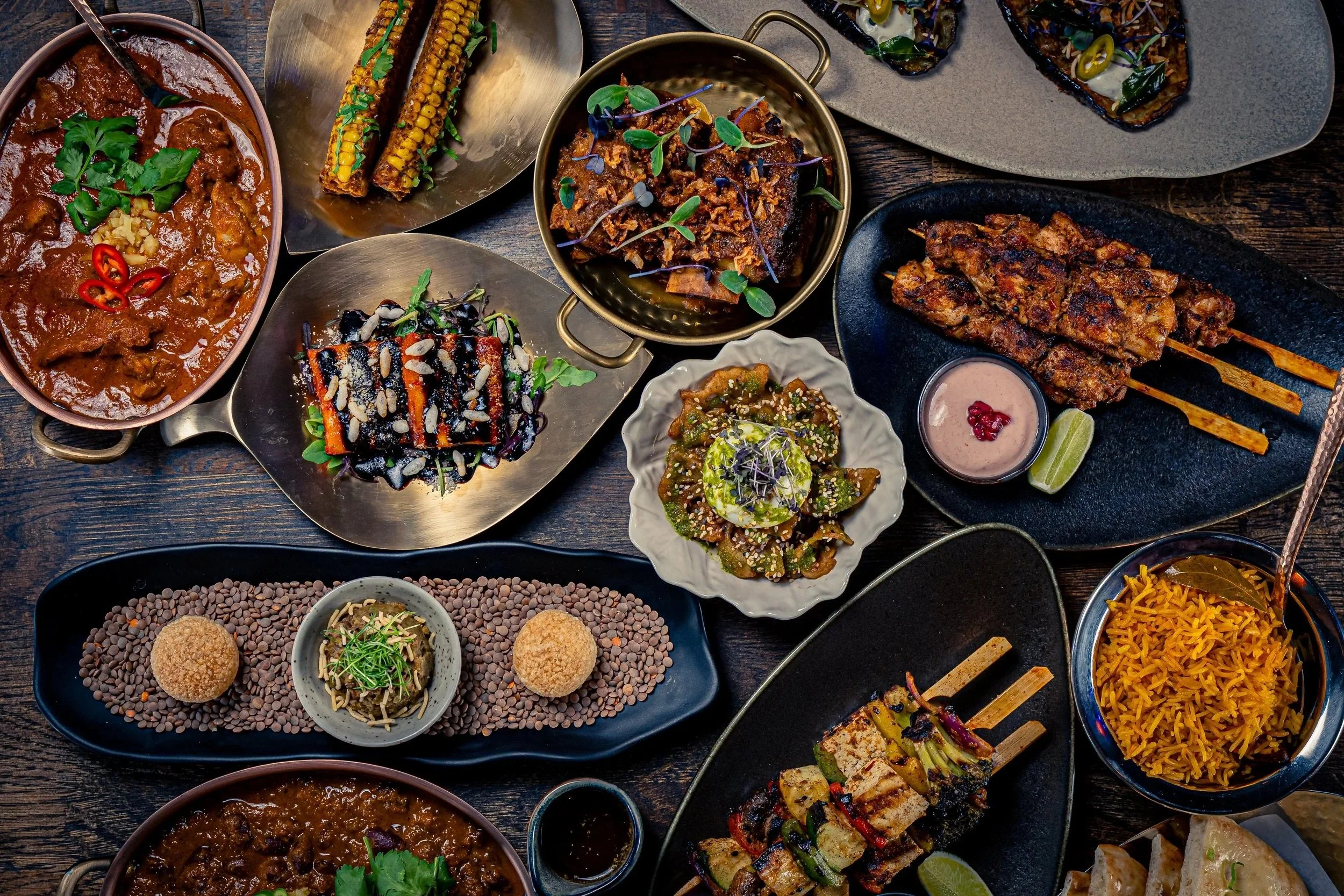 A variety of Indian dishes served on a dark wooden table, including curries, grilled skewers, rice, and garnished vegetables with lime wedges.