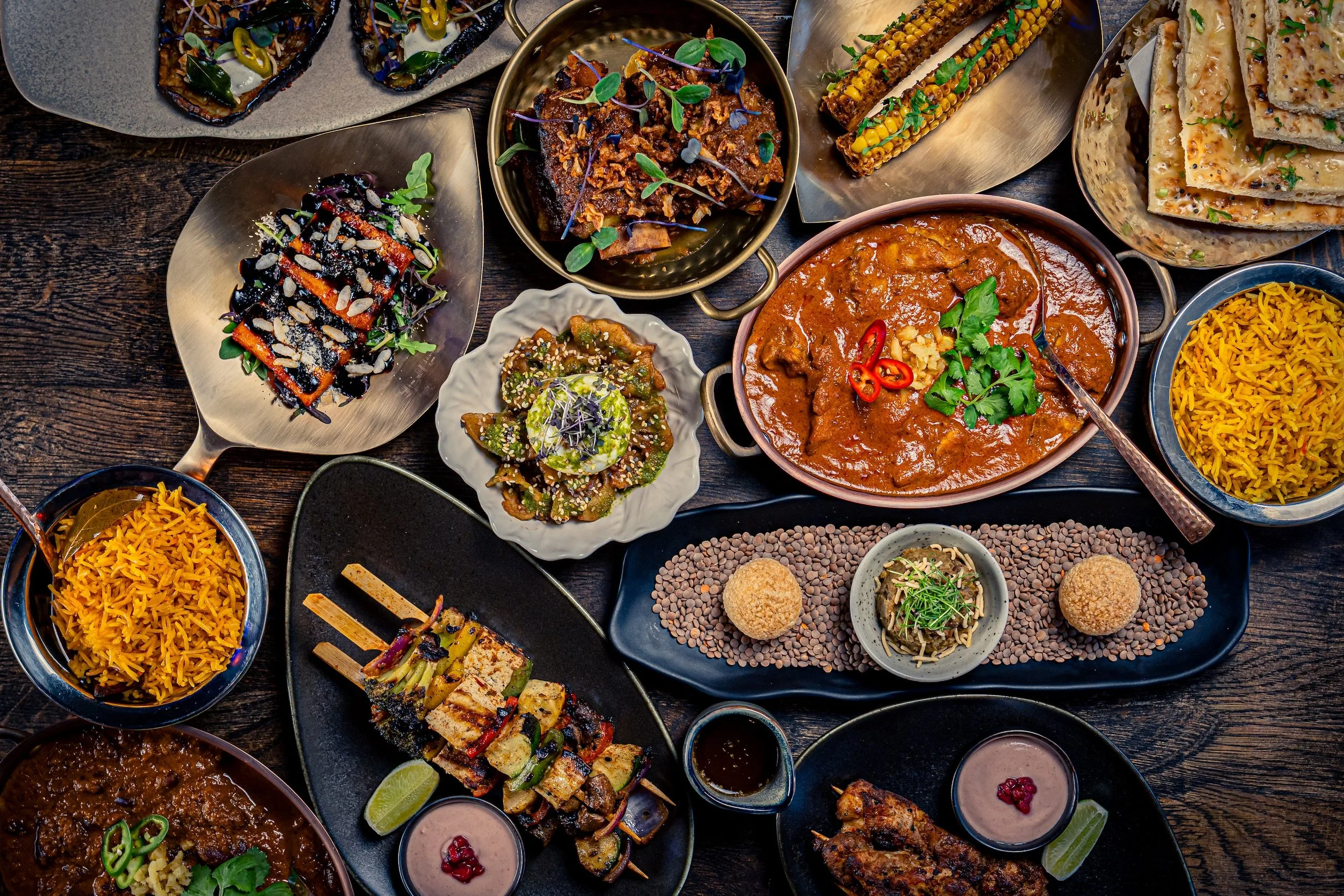 Assorted Indian dishes on a dark wooden table, including curries, rice, grilled skewers, stuffed bread, and small appetizers.