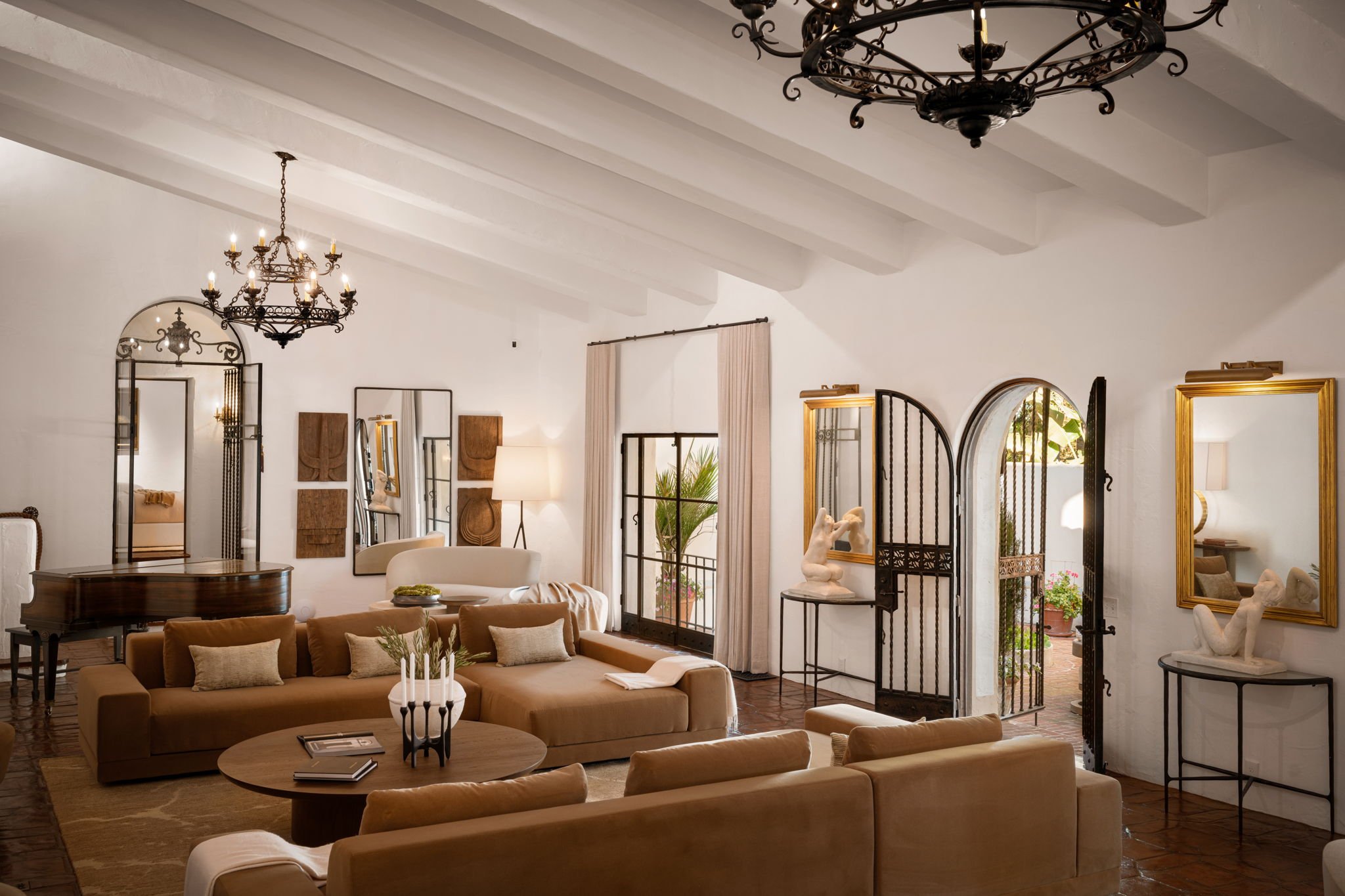 Living room with beige sofas, a round wooden coffee table with a plant and books, two windows with curtains, decorative sculptures, and an open black iron gate leading to an outdoor space with a fountain and brick walls.