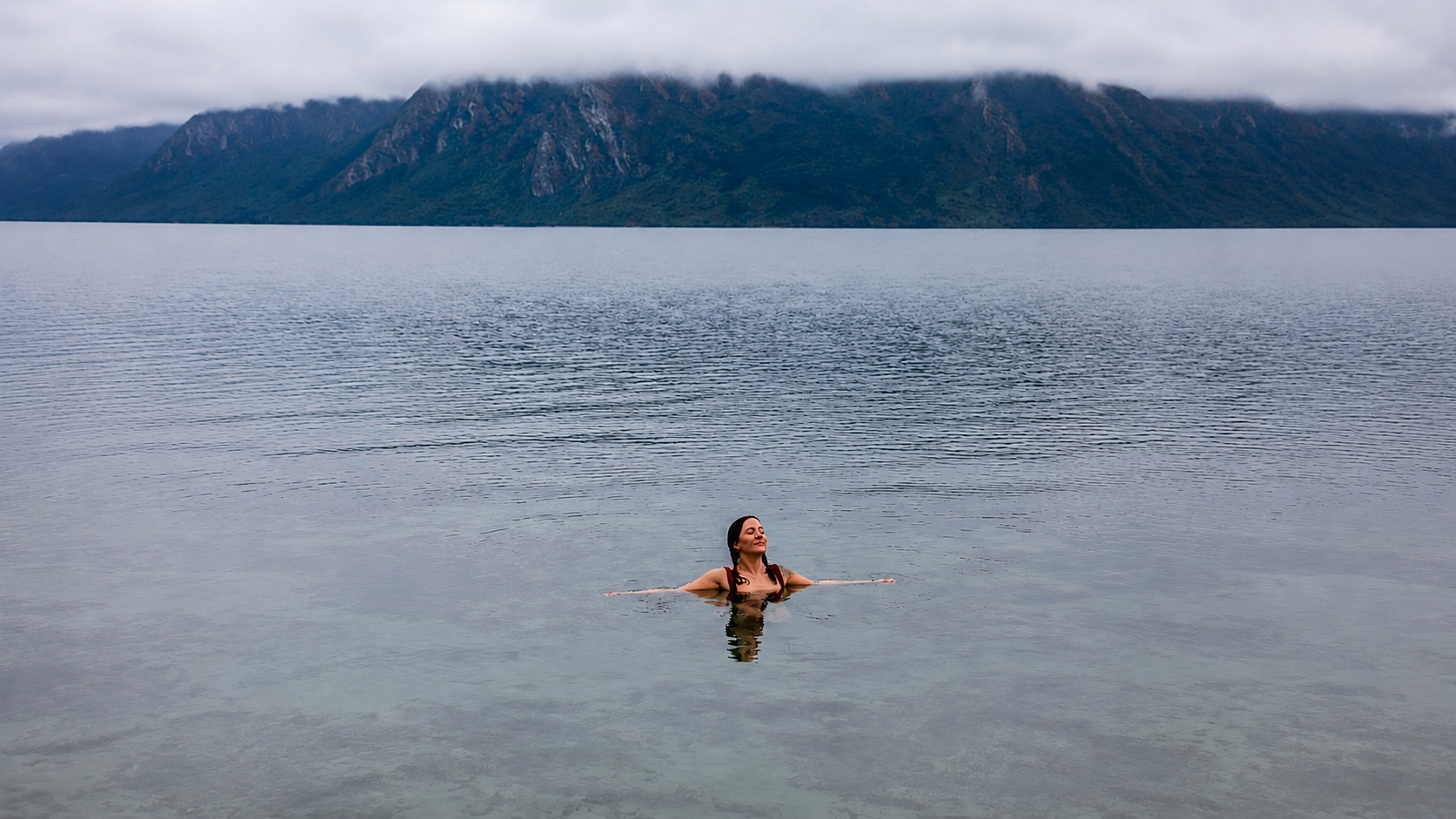 Woman relaxing in Lake Wānaka, surrounded by mist-covered mountains