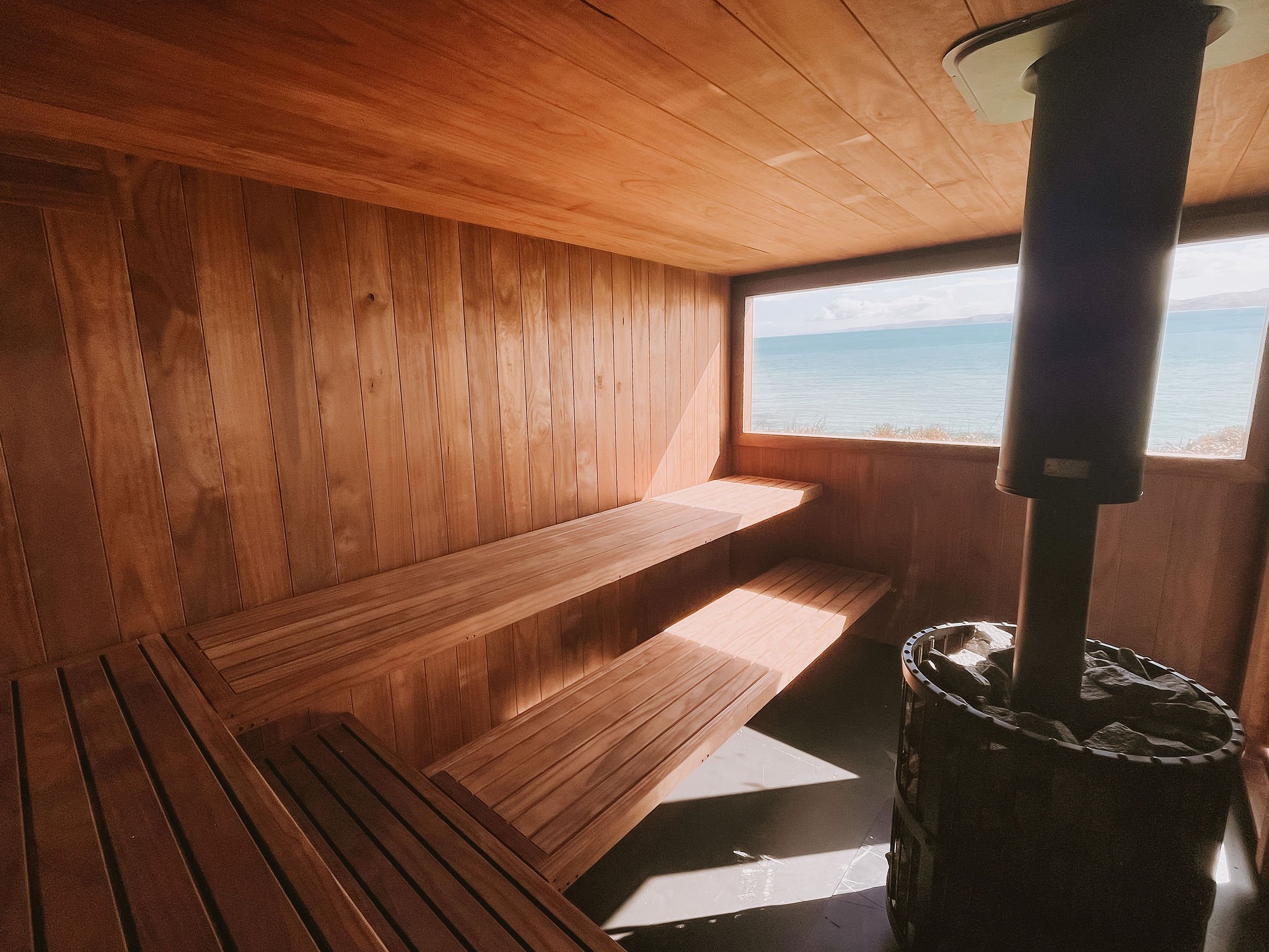 Interior of The Sauna Wānaka featuring natural timber benches, a traditional wood-fired sauna heater and panoramic lake and mountain views.
