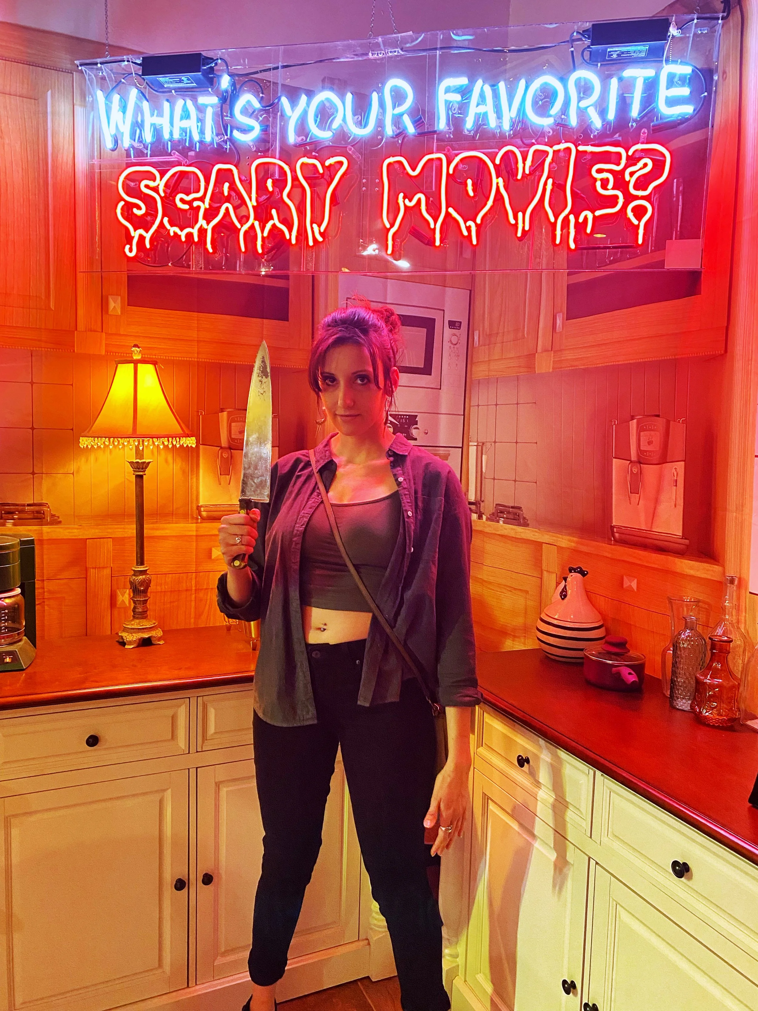 Chelsea poses with a fake knife in a kitchen that resembles the set from Scream with the words "What's your favorite scary movie?" above her