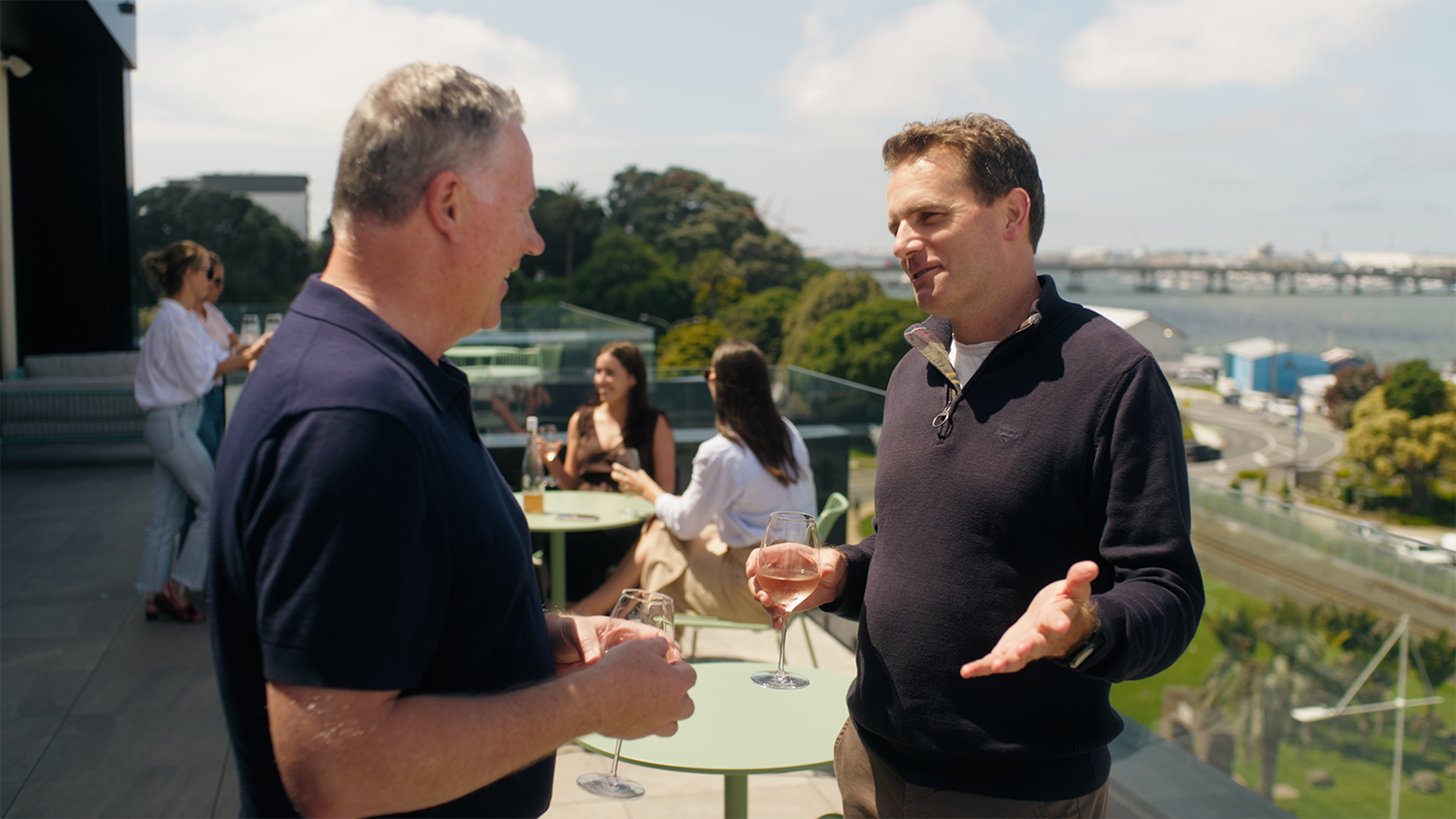 Two men having a conversation with glasses of rosé on a balcony overlooking a city and water, with three women sitting nearby in the background.