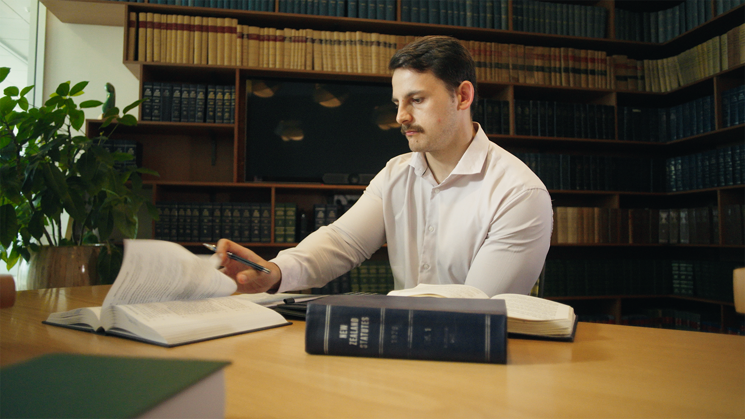 A man with dark hair and a mustache sits at a wooden table in a library, surrounded by bookshelves filled with law books, as he studies and reviews documents.