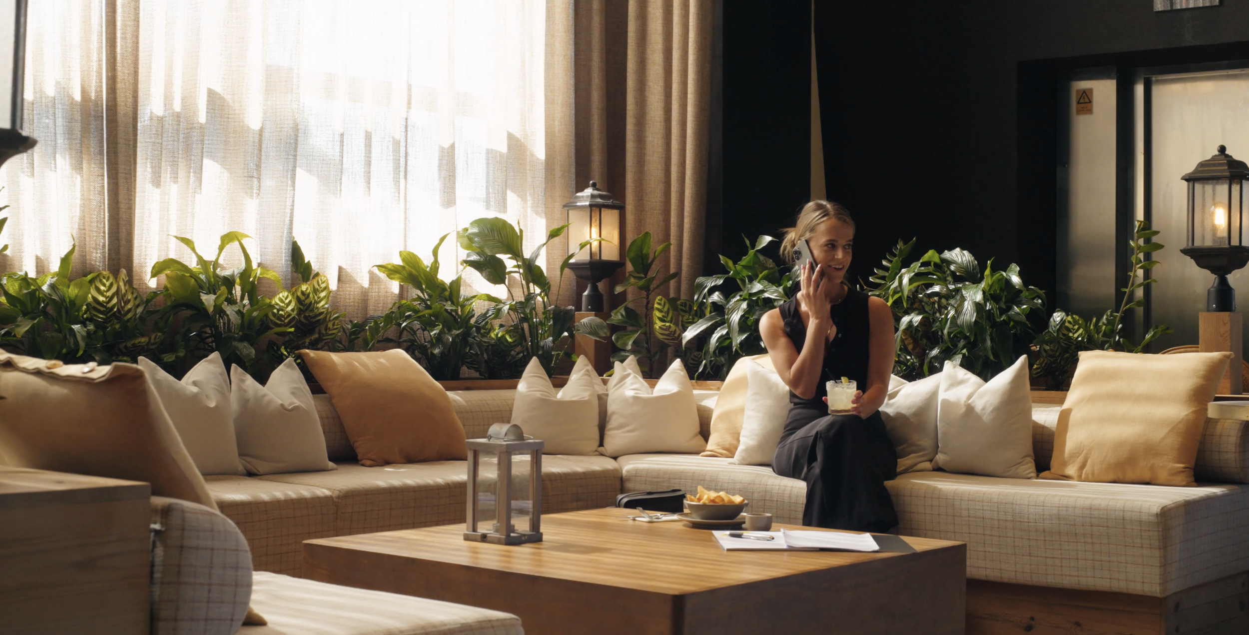 A woman in a black dress sitting on a beige sofa with white and yellow pillows, talking on a phone, holding a drink, in a cozy room with curtains, plants, and lamps.