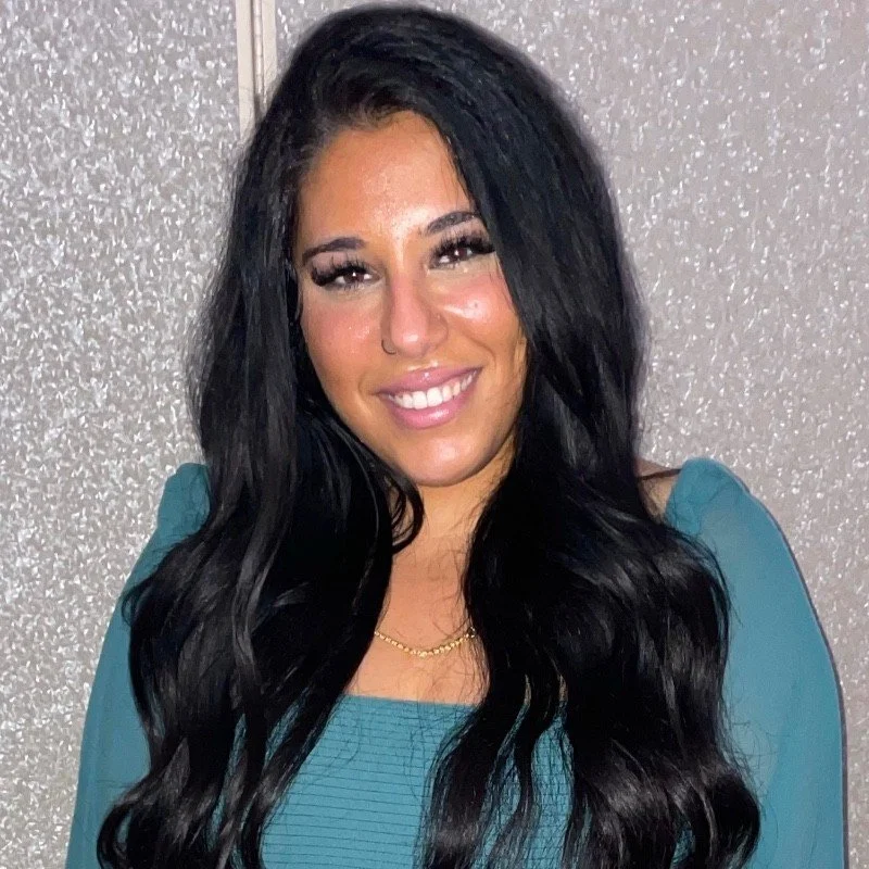 A young woman with long black hair, smiling, wearing a teal top and a gold necklace, standing against a sparkly silver background.