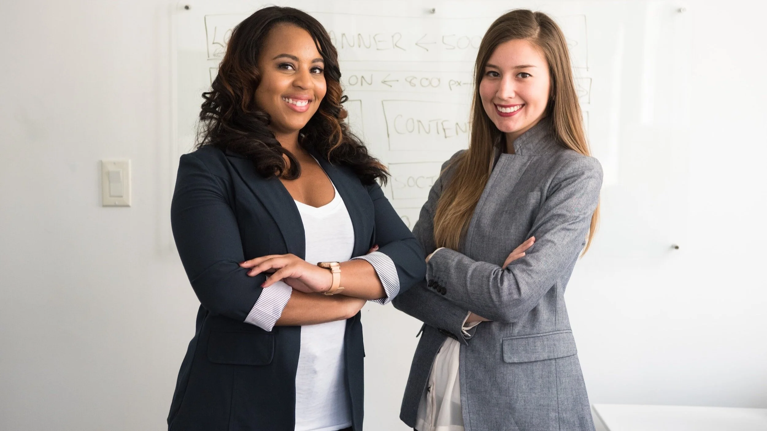 Two women standing together in an office or conference room, smiling at the camera, with a whiteboard in the background.
