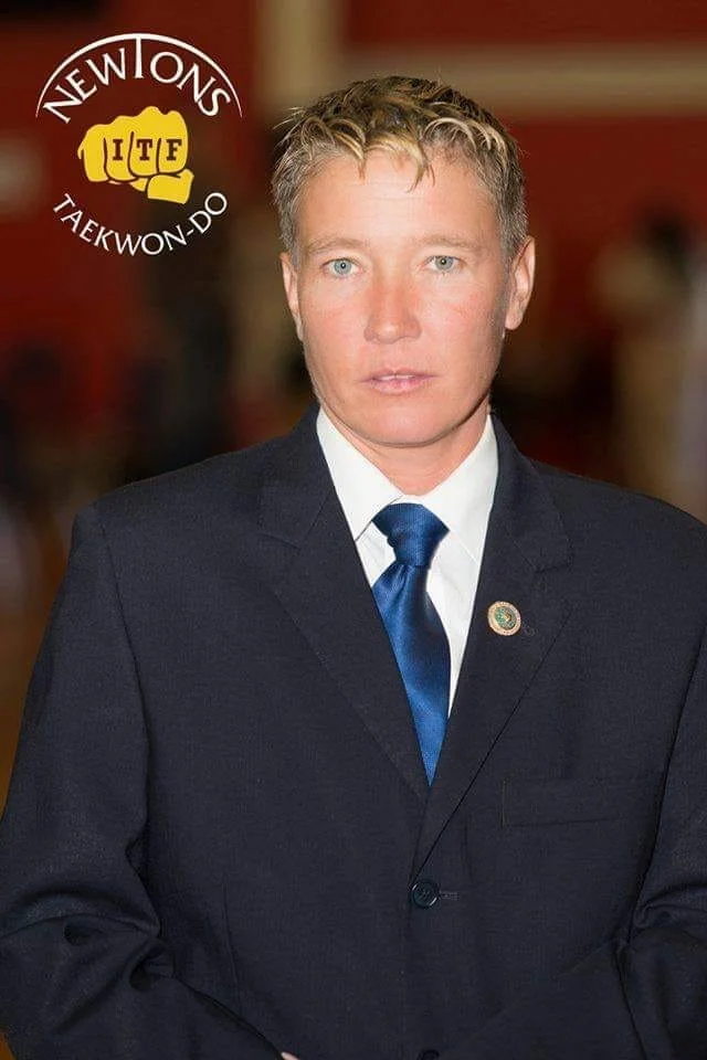 A man in a suit with a blue tie standing indoors. There is a logo in the top left corner that says 'NEW OMN' and 'TAEKWON-DO' with a yellow fist and 'ITF' in red letters.