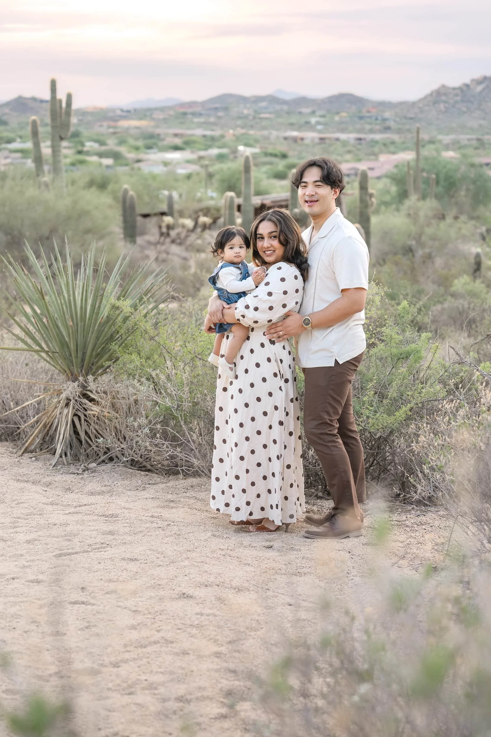 A family of three, including a woman, a man, and a young girl, standing outdoors in Scottsdale Arizona's desert landscape with cacti and mountains in the background, during sunset.