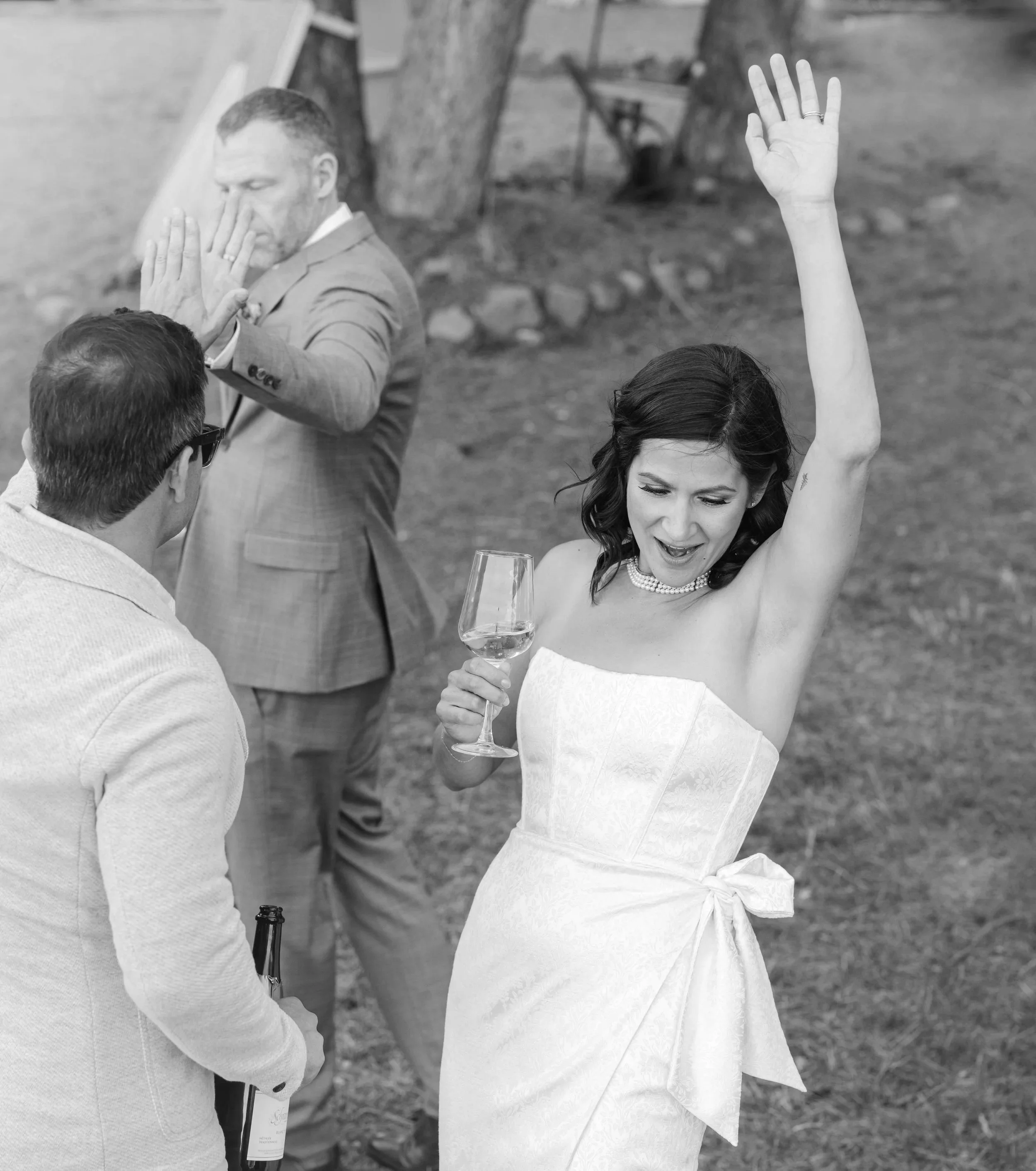 Emotive scene with a bride celebrating after her ceremony, arm raised with a glass of Champagne. Groom is giving his best man a high five.