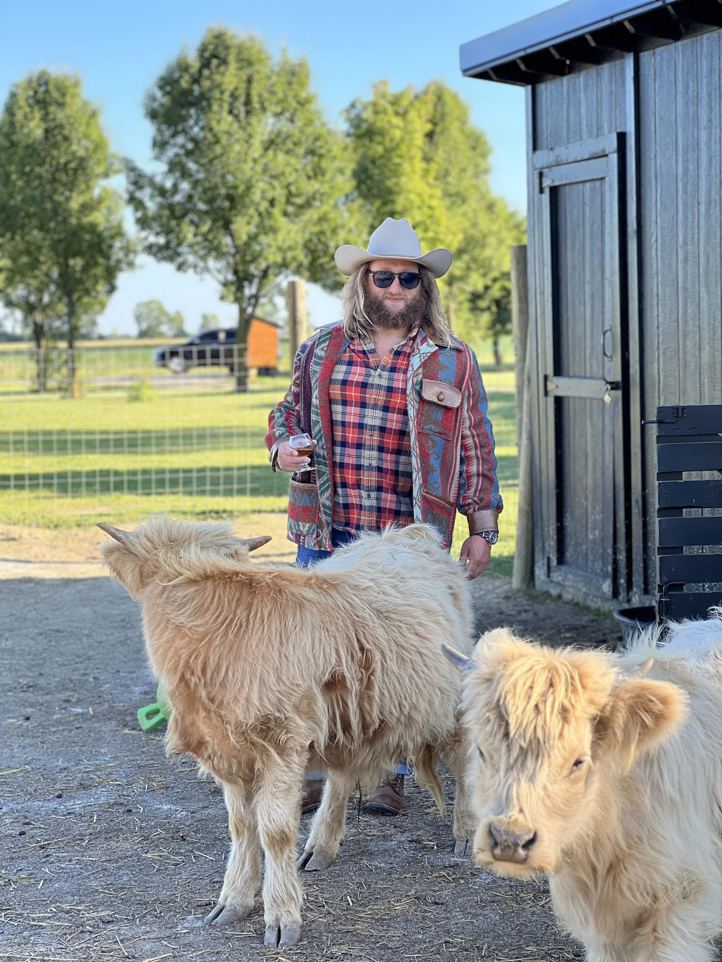 A man with long hair, beard, wearing sunglasses, a cowboy hat, a colorful plaid shirt, and a patterned jacket, holds a glass of wine standing outdoors next to a wooden structure with baby goats nearby.