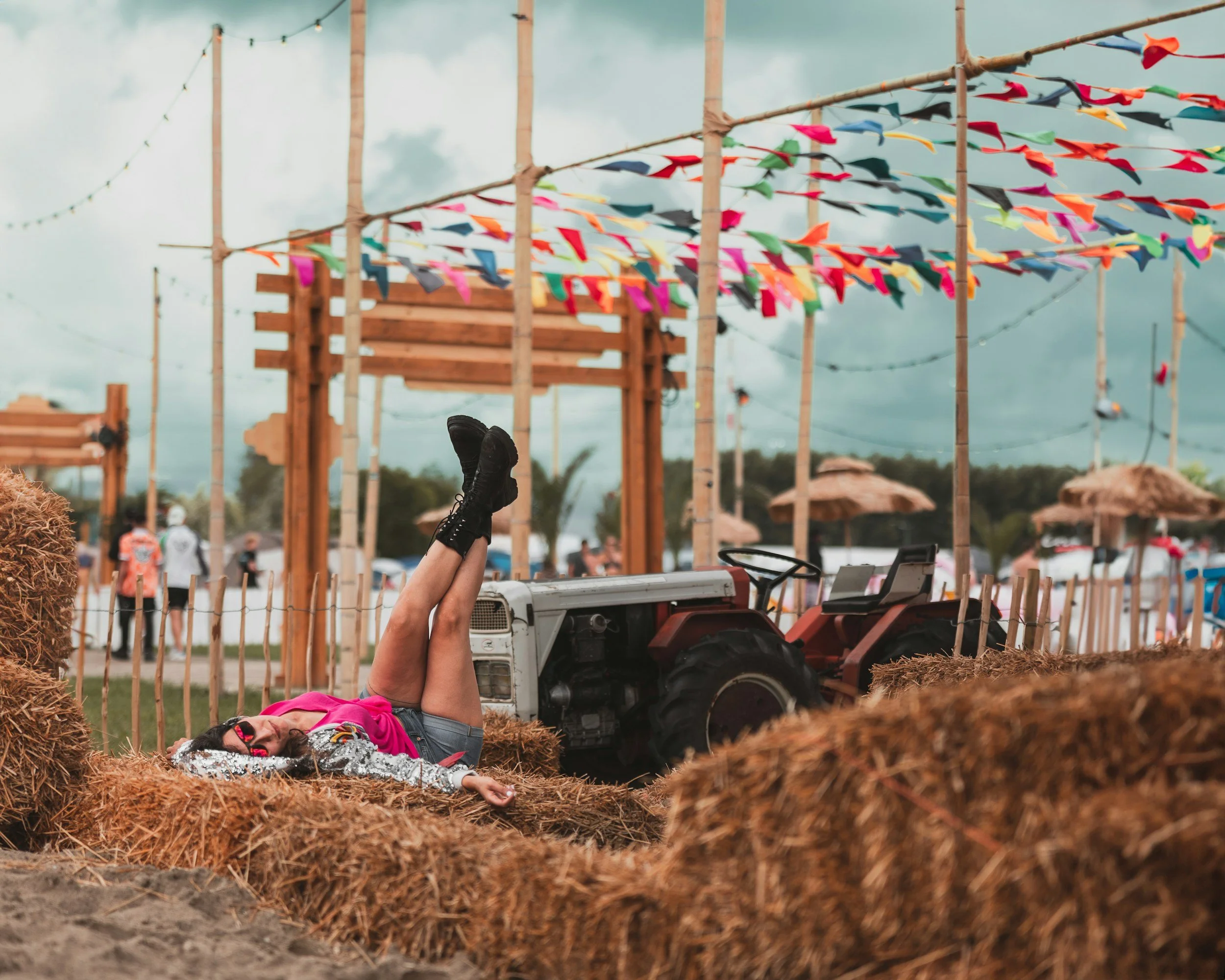 A person lying on a hay bale with legs up, wearing sunglasses, a pink shirt, and shorts, at an outdoor festival with colorful banners, a tractor, and umbrellas in the background.