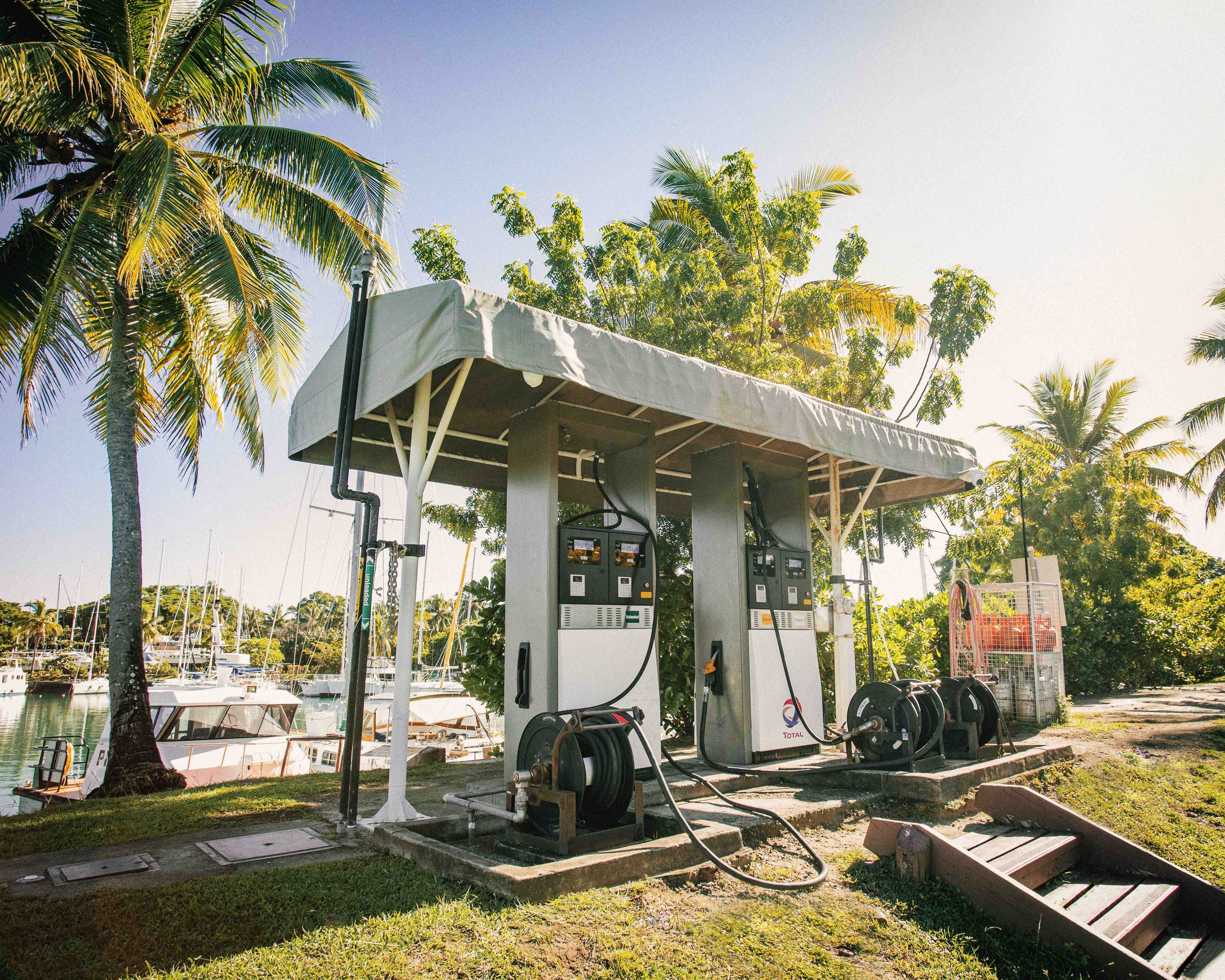 Outdoor gas station near a marina with boats and palm trees.