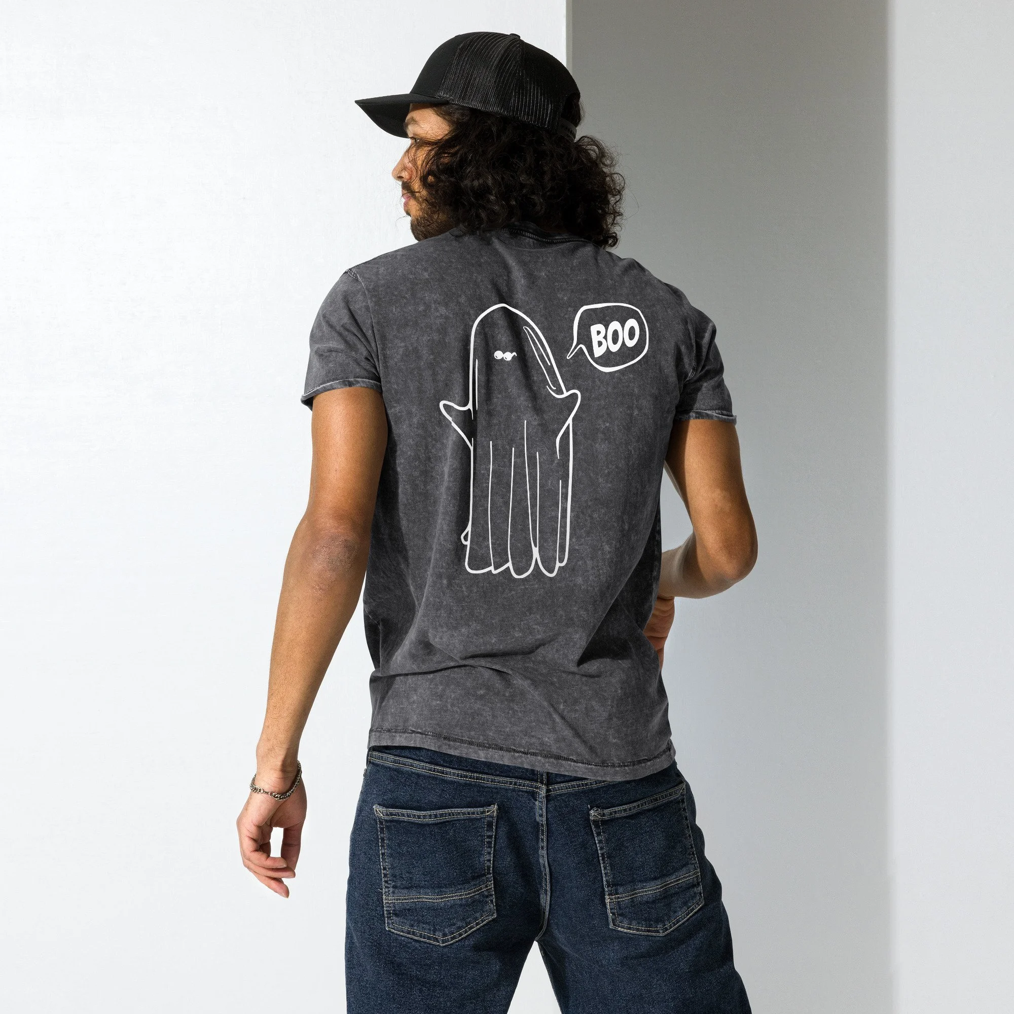 A man wearing a black cap and a Pedestrian brand grey t-shirt with a white outline of a ghost saying 'BOO' on the back, standing in front of a plain white and gray wall.