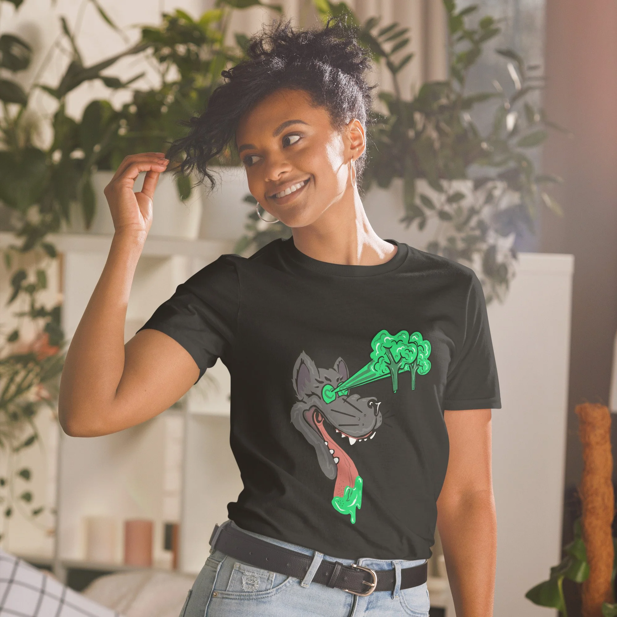 A woman with curly black hair wearing a Pedestrian brand black t-shirt featuring a graphic of a smiling drooling wolf with laser-like stream of broccoli from its eye, standing indoors with plants in the background.