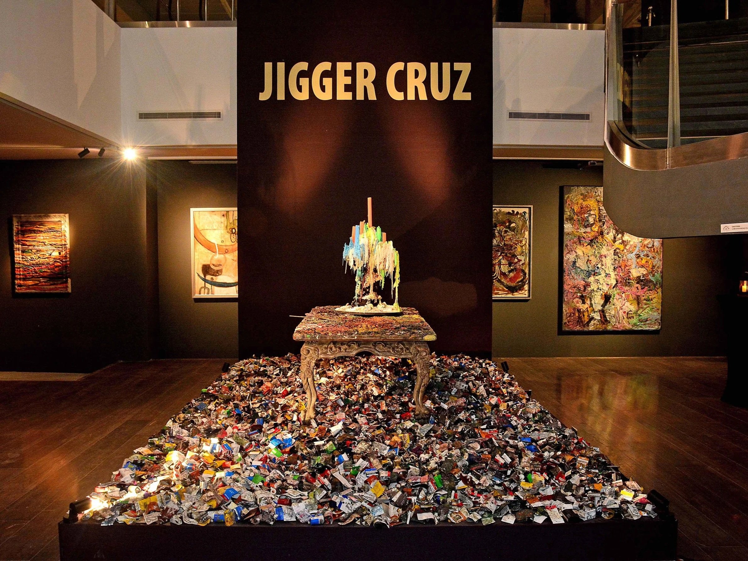 Art exhibit in a gallery with a pedestal covered in colorful candy wrappers rained on the floor, a dark background wall with the text 'Jigger Cruz' and a mixed media sculpture on top, surrounded by abstract paintings.