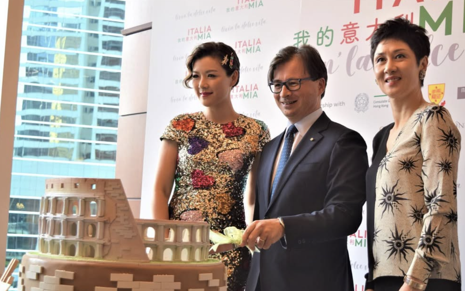 Three people, two women and one man, are gathered around a cake during a celebration. The background features a white banner with red and green text and logos. The woman on the left wears a colorful, floral-patterned dress, the man in the middle is i