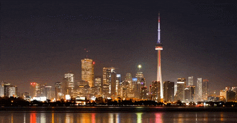 Nighttime city skyline with illuminated skyscrapers and a tall tower reflected on water.