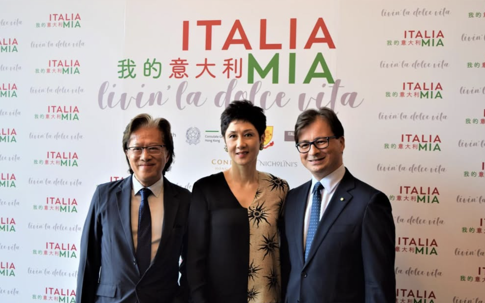 Three people in formal attire standing in front of a backdrop with Italian and Chinese writing, smiling at the camera.