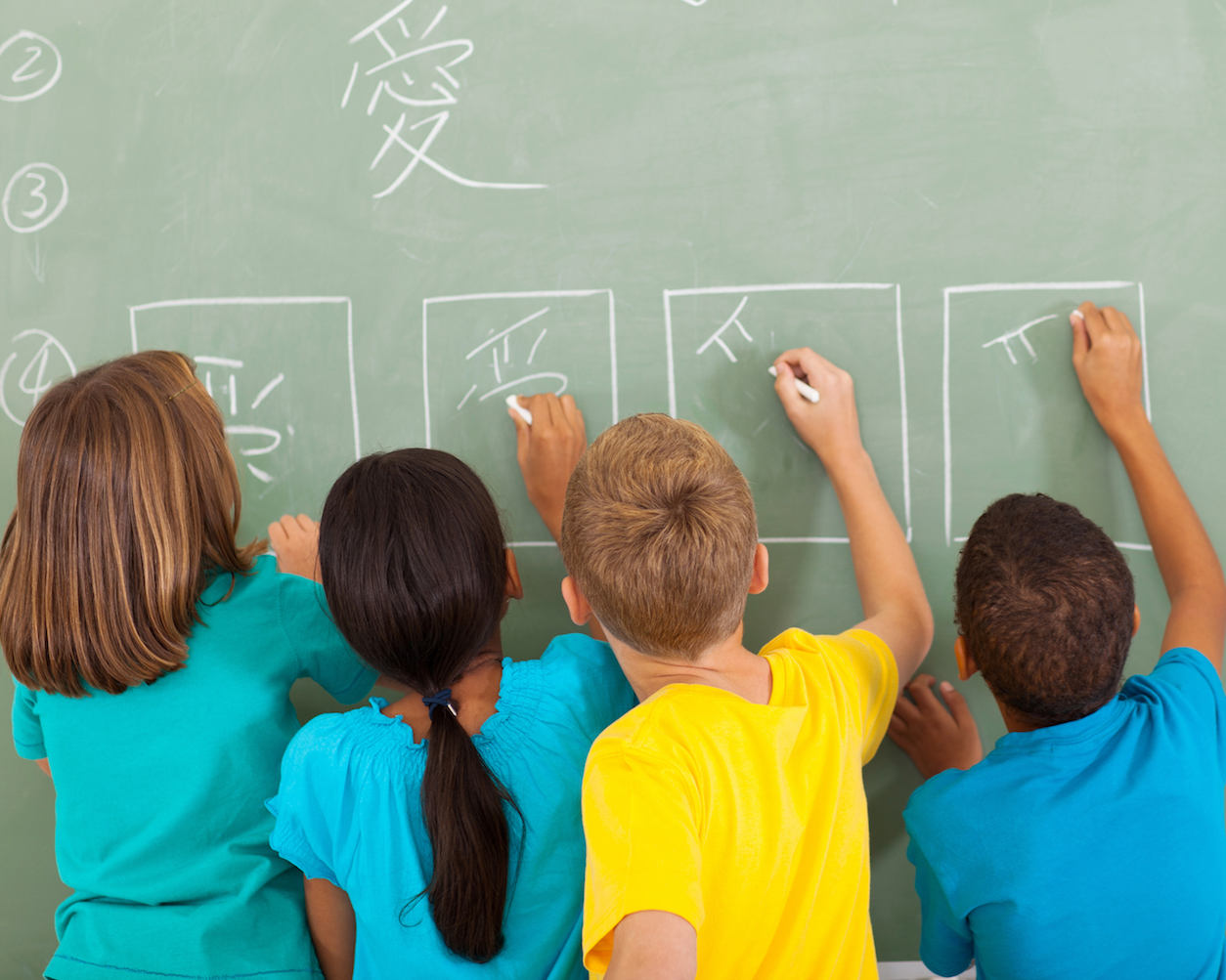Four children writing Chinese characters on a green chalkboard with chalk, viewed from behind.