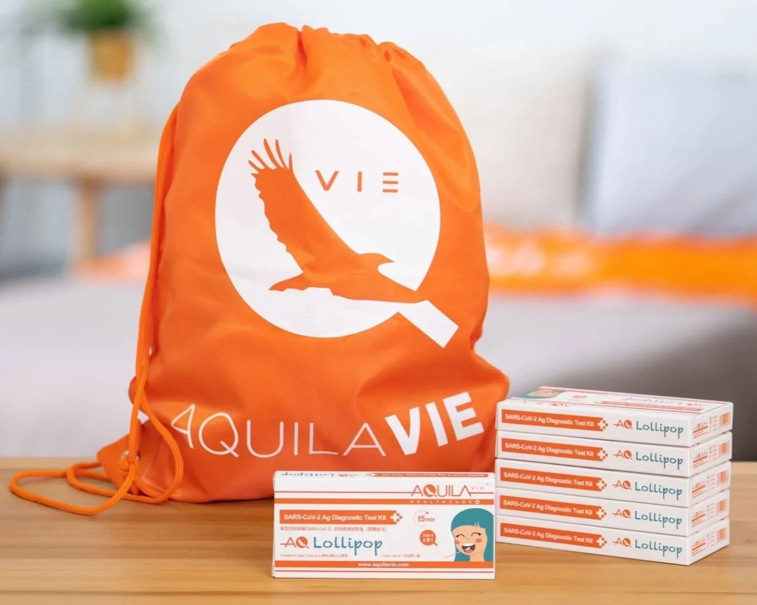 Orange reusable bag with a white logo of a bird and the text 'AQUA VIE' on a wooden table. In front, there are five boxes of SARS-CoV-2 antigen test kits, each labeled 'AQ Lollipop' with a smiling face illustration, and one additional test kit box positioned horizontally.