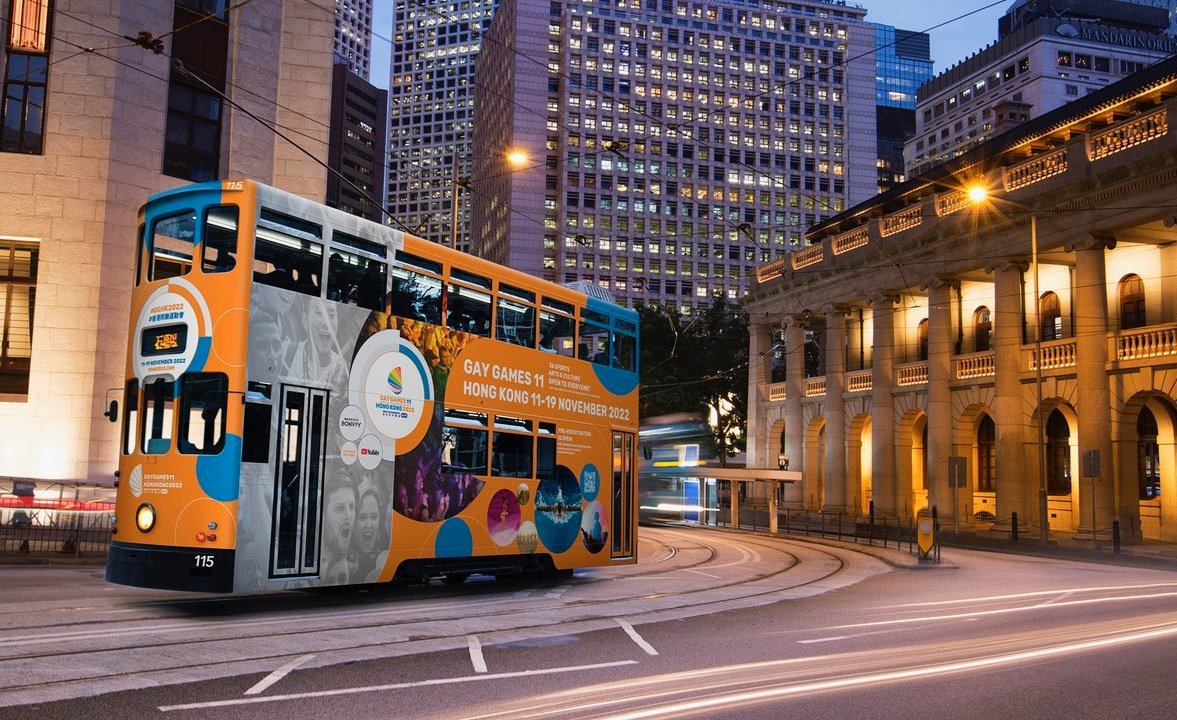 A double-decker tram decorated with advertisements for the Gay Games 11 in Hong Kong, traveling on city streets at dusk with tall buildings and historic architecture illuminated in the background.