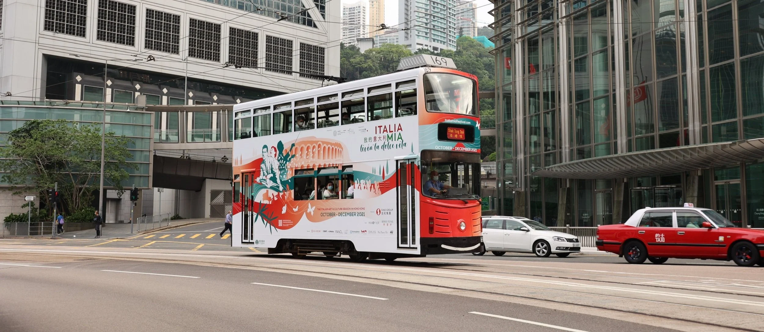 Double-decker tram with colorful art and text advertising Italian culture festival in Hong Kong, moving along a city street with office buildings and cars around.
