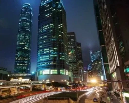 Night view of illuminated skyscrapers in a city, with streaks of car lights on the busy street below.