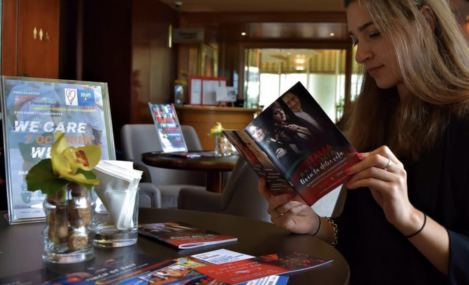 A woman with long light brown hair reading a brochure or flyer at a table in a cozy, wood-paneled indoor setting, with promotional posters and flower vases on the table.