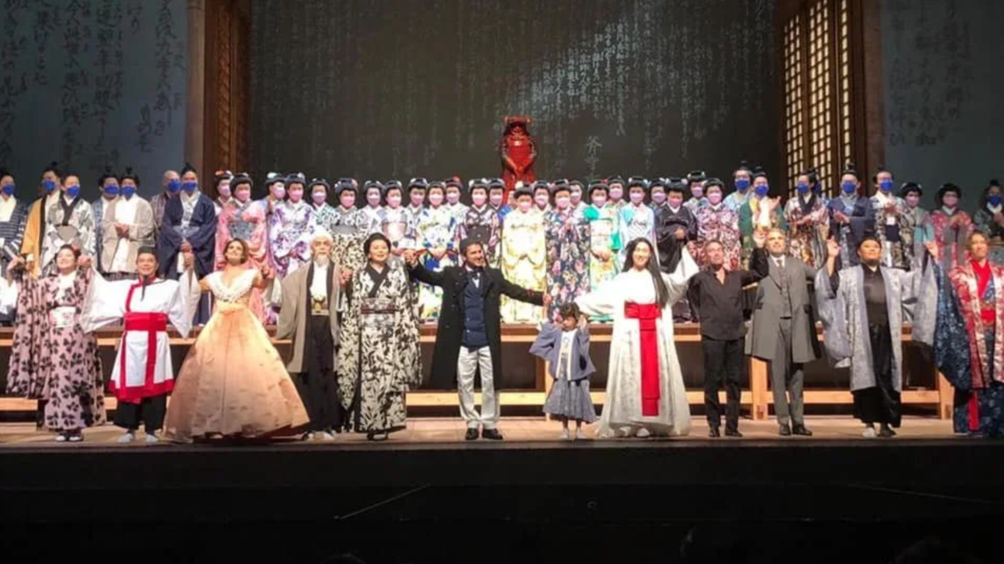 Group of people wearing traditional Japanese kimonos and masks, standing on a stage, likely after a cultural performance or event, with a backdrop featuring Japanese calligraphy.