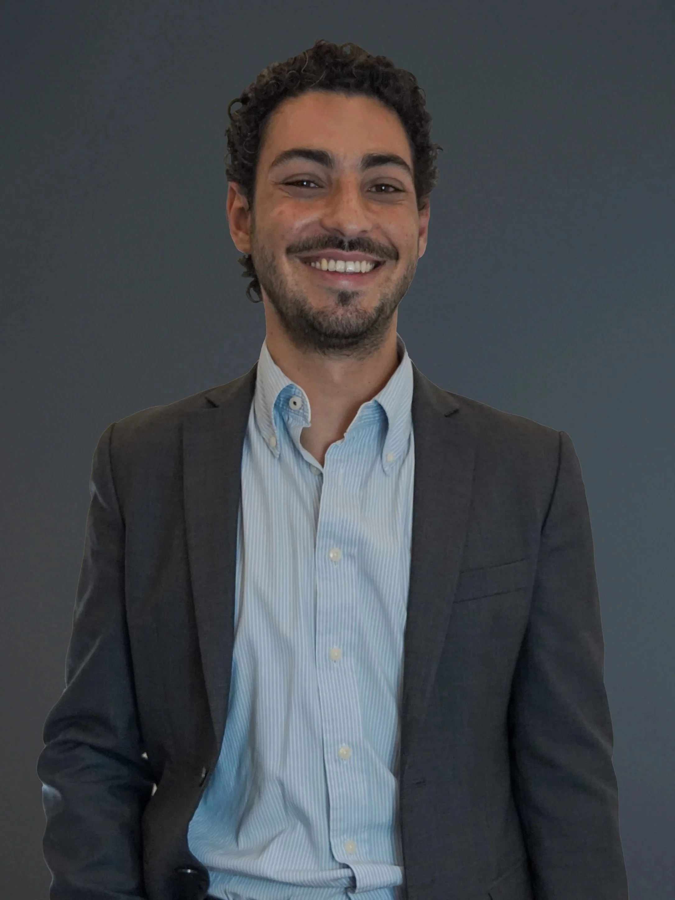 Smiling man with dark, curly hair and a beard wearing a gray suit and light blue dress shirt, standing against a dark background.