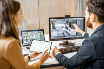 Two people in a meeting room with laptops and a desktop monitor displaying architectural designs.