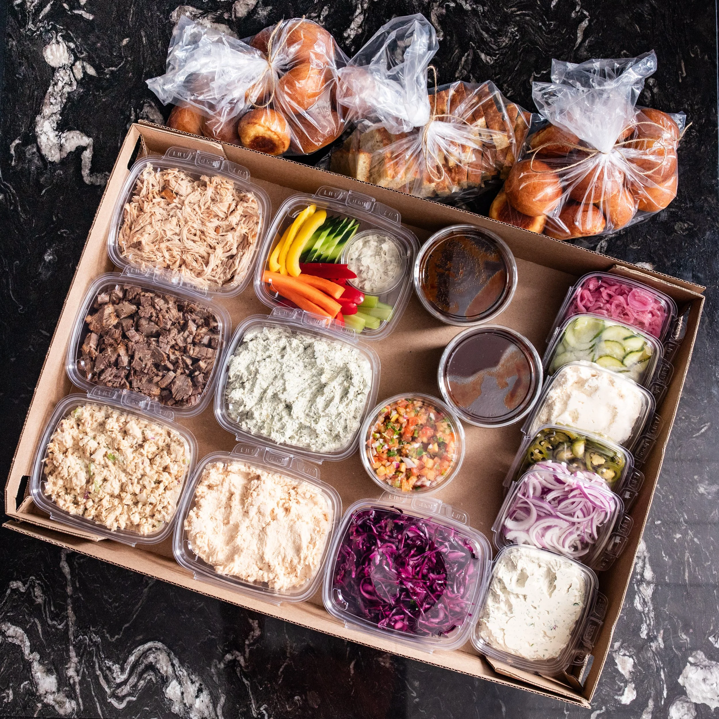 A cardboard box with assorted containers of dips, pulled pork, chicken salad, brisket, and condiments, with bags of sandwich bread and rolls for sliders on a dark marble surface.