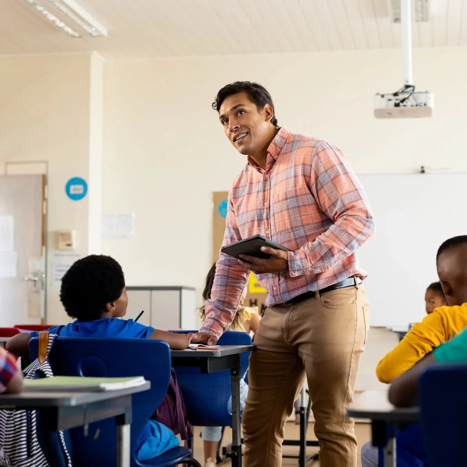 Young male middle school teacher in classroom with tablet, standing at student's desk