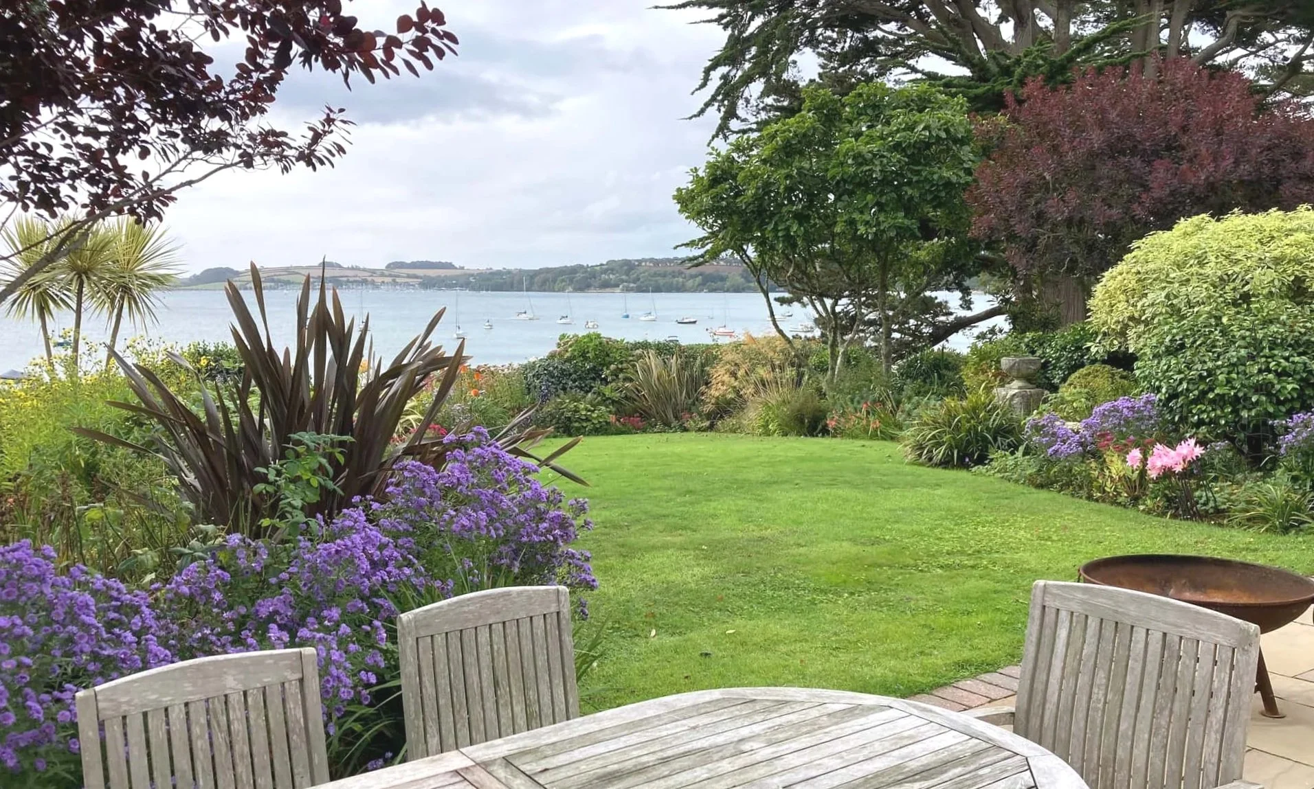Teak furniture, a fire bowl, and a beautifully kept estaury garden with asters and nerines in flowerr, boats on the estuary