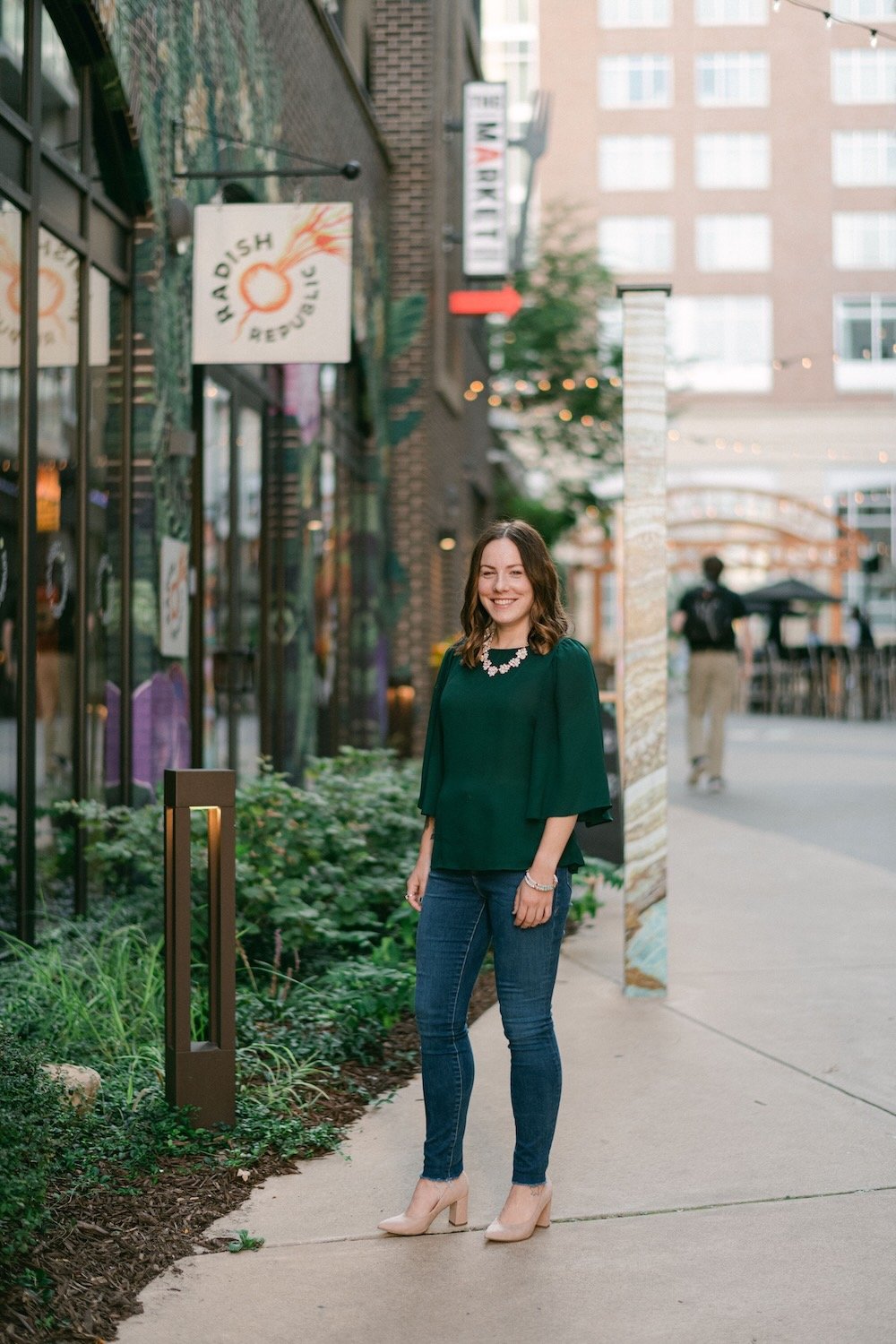 A woman standing on a sidewalk outside a restaurant with a sign that says 'Har-dish Beverages', smiling, wearing a green top, jeans, beige high heels, and a necklace.