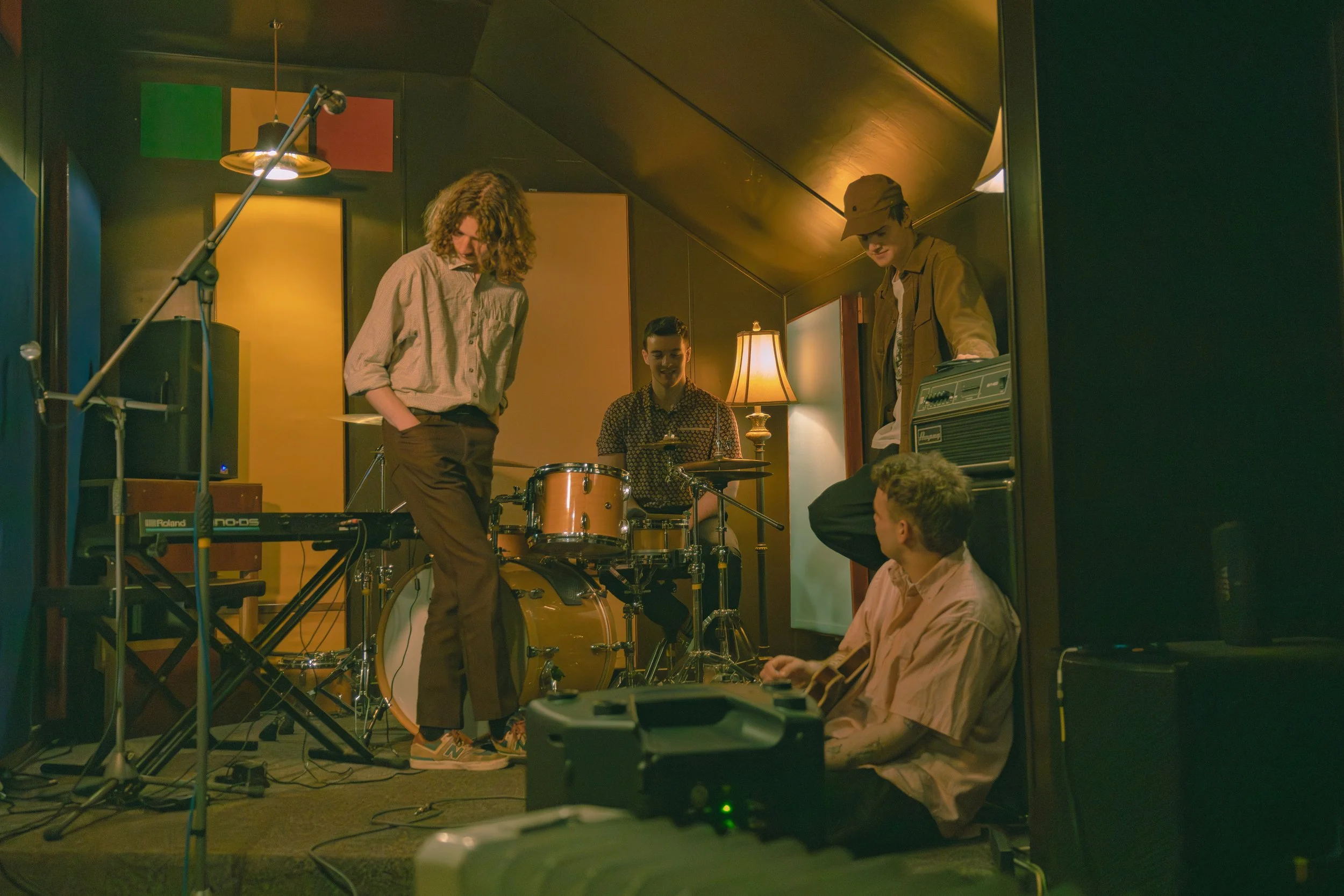 A group of young people in a recording studio with musical instruments, microphones, and audio equipment, some sitting and some standing, engaged in a musical activity.