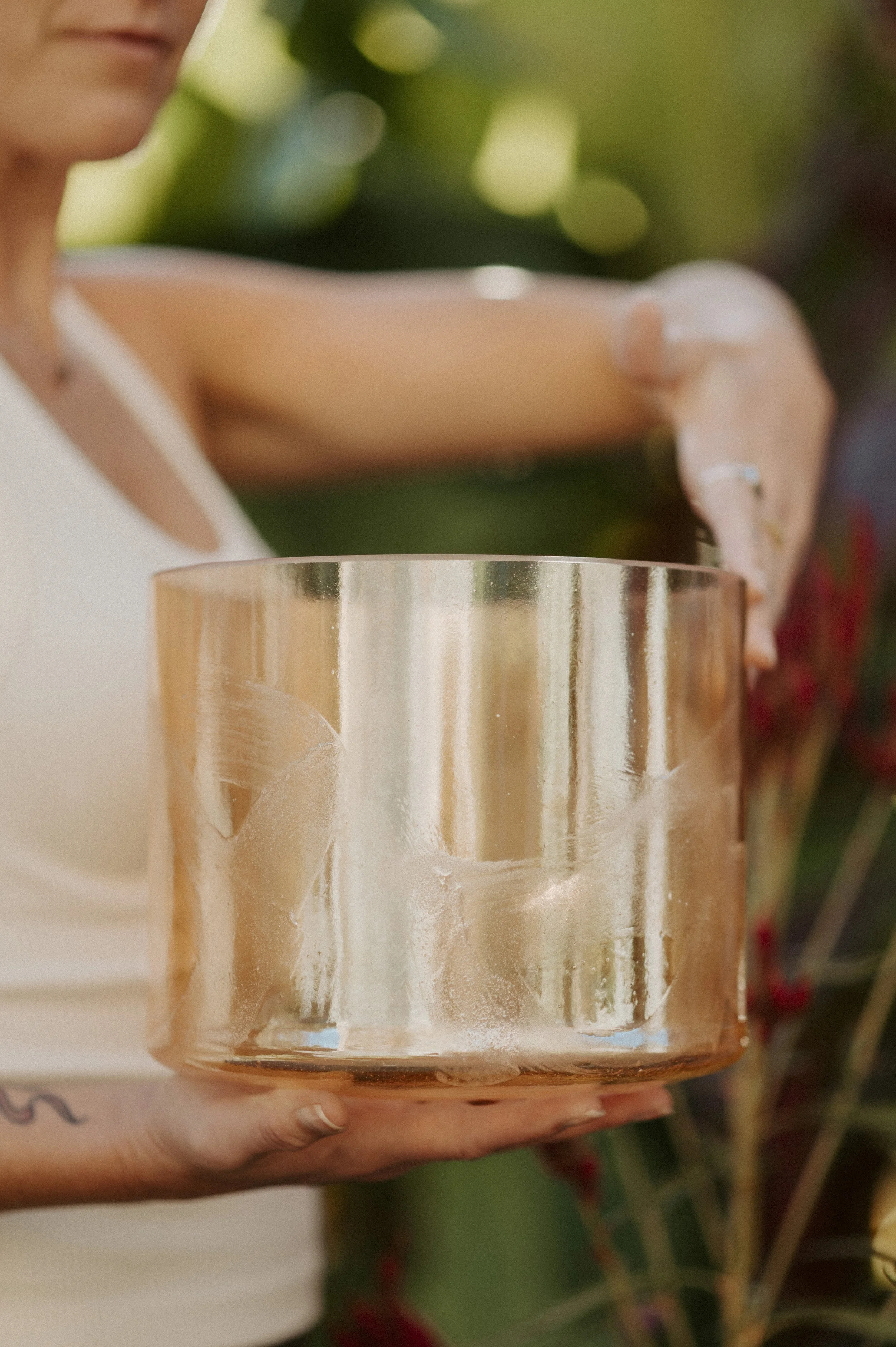 A woman holding a large, shiny, orange-colored singing bowl outdoors during the daytime, with greenery in the background.