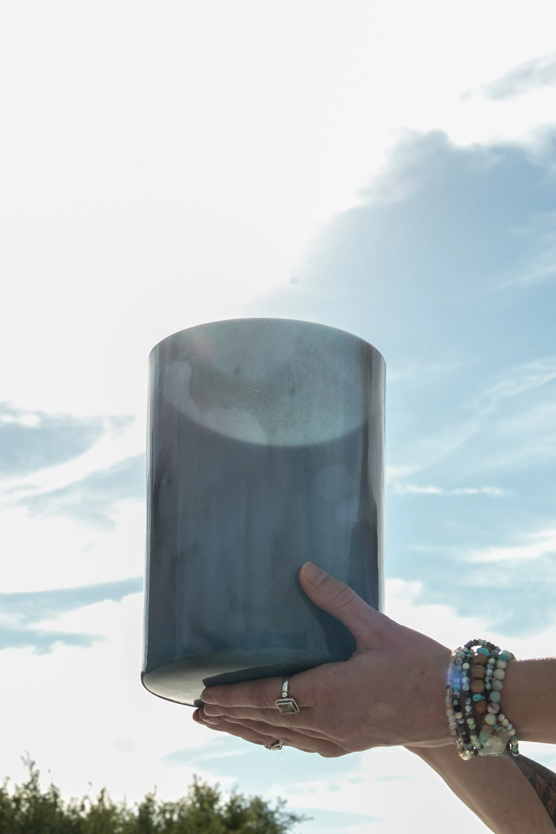 A person’s hand with multiple bracelets and a ring on the finger holding a large blue Crystal Tones singing bowl outdoors against a partly cloudy sky.