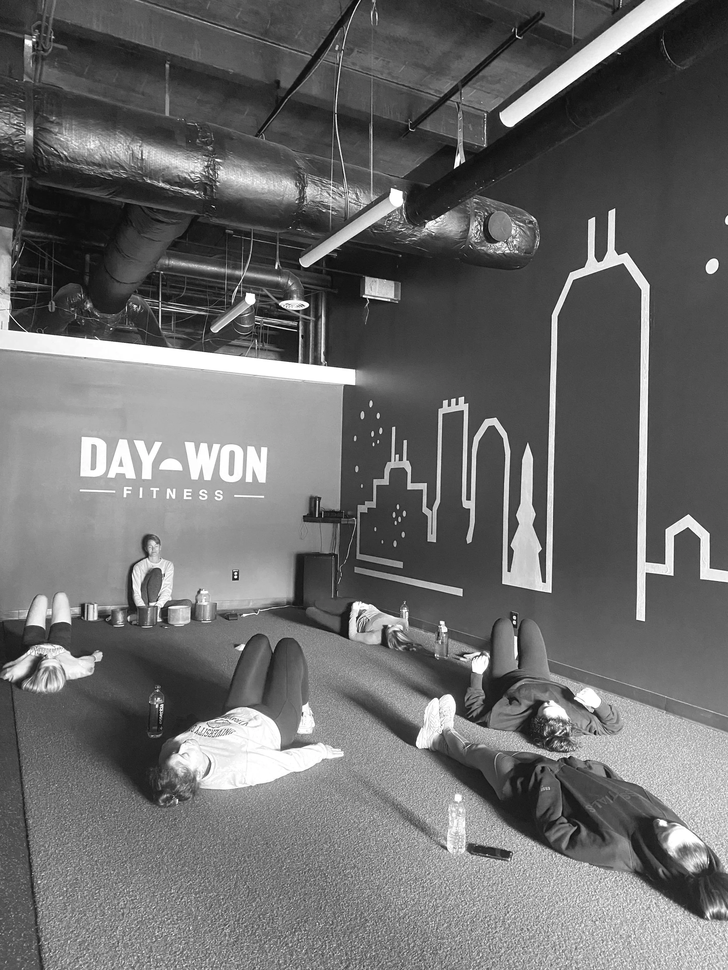 Athletes lying on the artificial turf floor of Day Won Fitness, a fitness studio, participating in a sound bath with water bottles and personal items nearby and a cityscape mural on the wall.