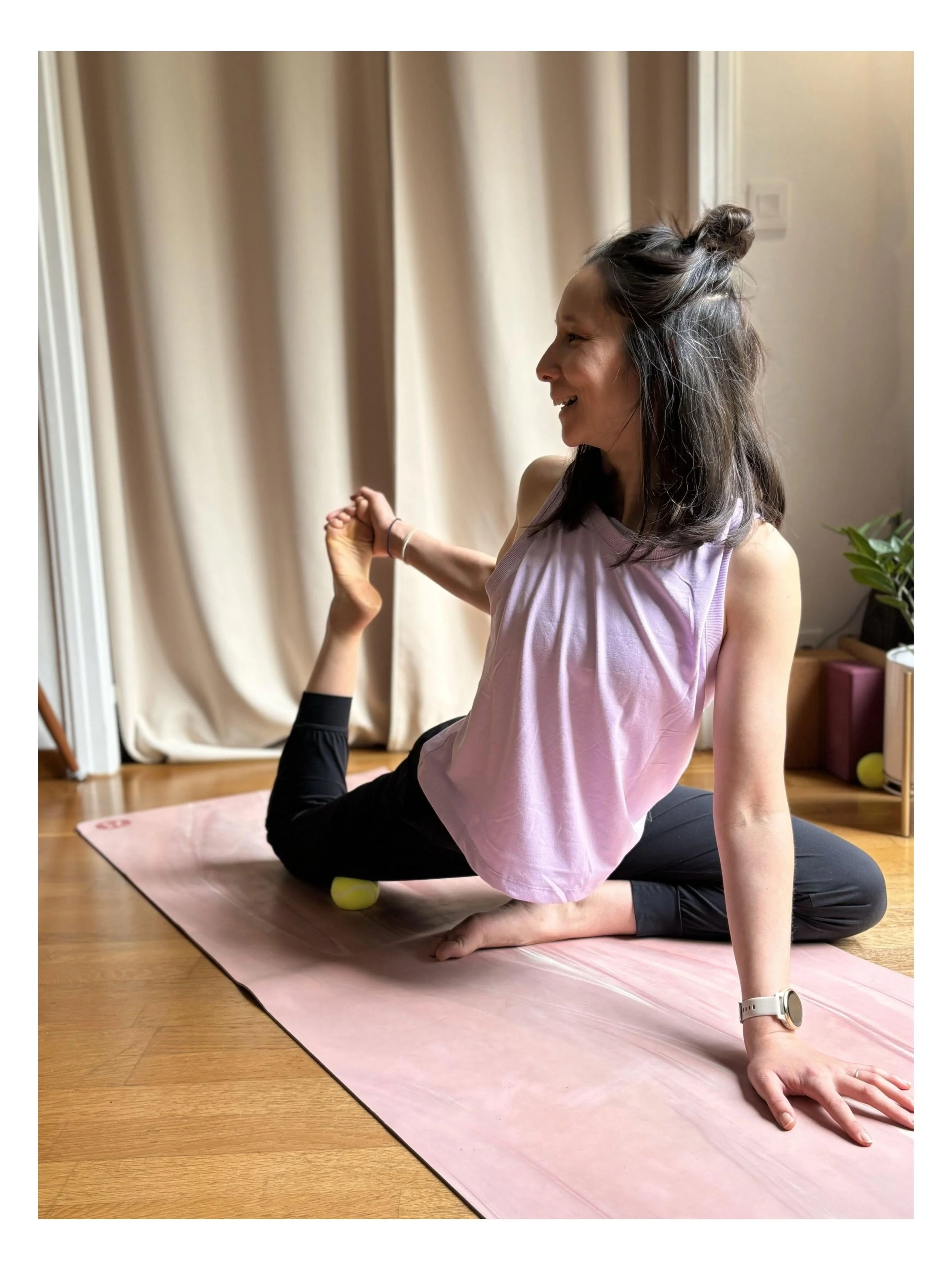 Une femme faisant du yoga ou des étirements à la maison, assise sur un tapis rose, vêtements de sport rose et noir, cheveux attachés en bun, dans une pièce avec des rideaux beiges, un mur clair, une plante verte et des meubles en bois.