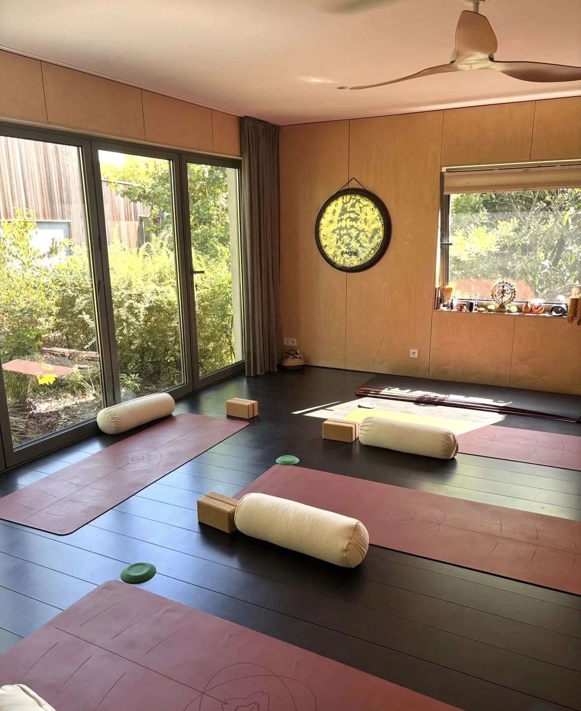 Studio27 Garches - Salle de yoga lumineuse avec des tapis de yoga rose, des blocs en bois, des cylindres de soutien et des coussins, face à de grandes fenêtres donnant sur un jardin vert.