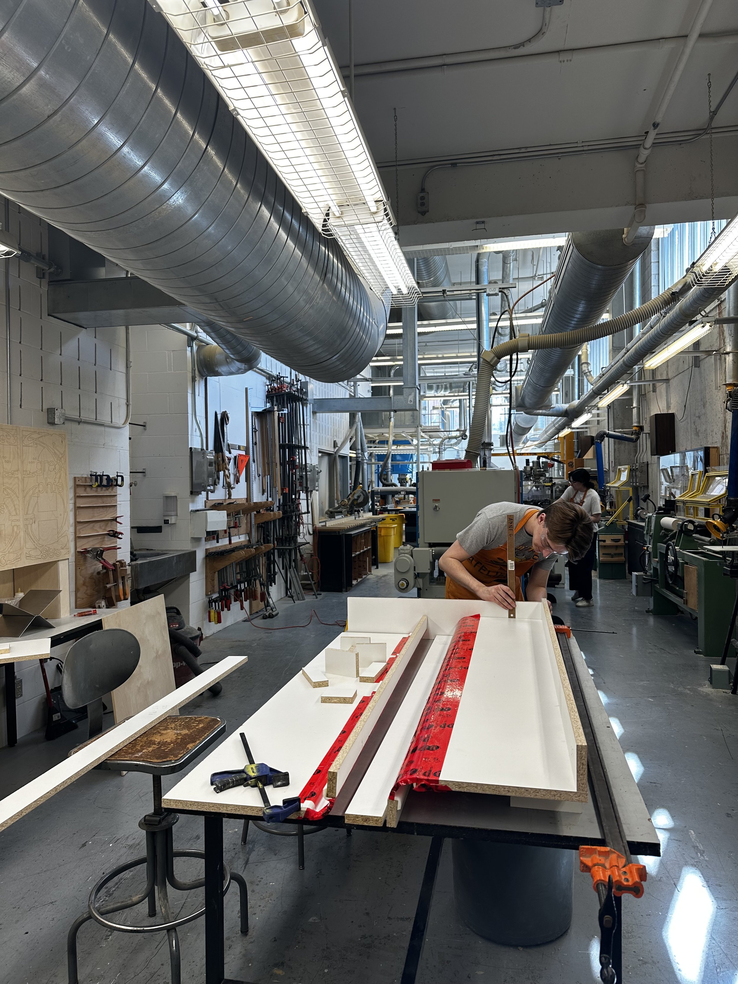 Une personne travaille sur une table de menuiserie dans une atelier de fabrication, avec divers outils et pièces de bois autour.