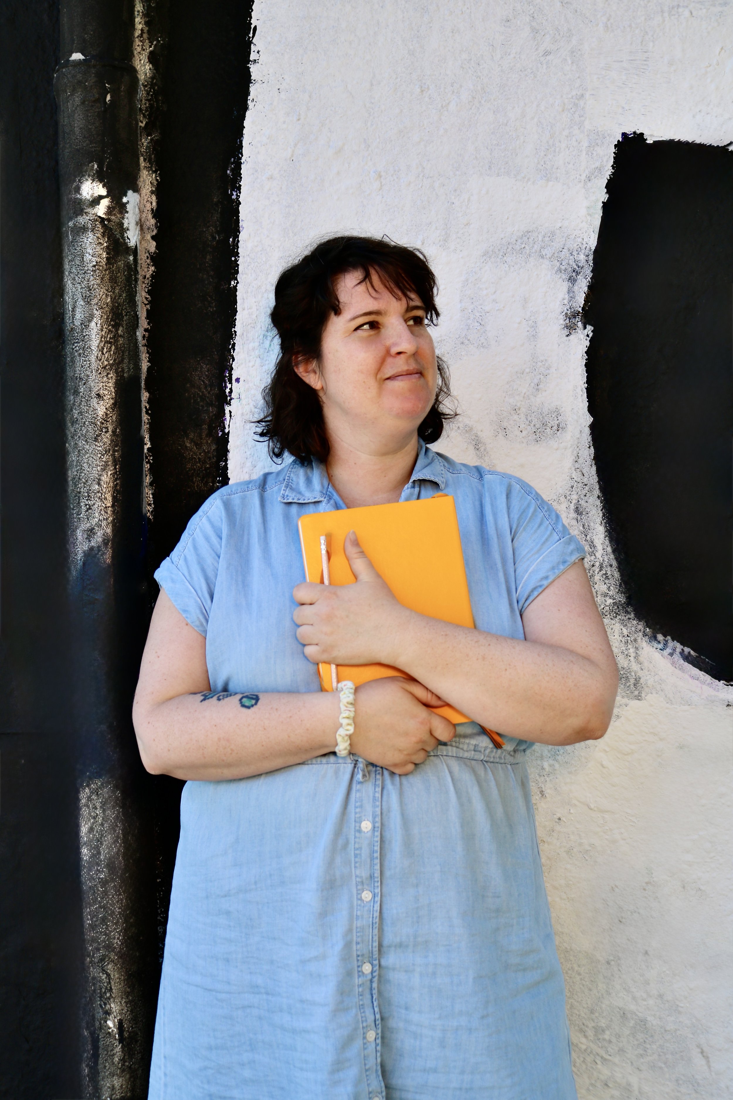 Sonja stands before a black and white wall with a bright orange sketchbook in her hands.